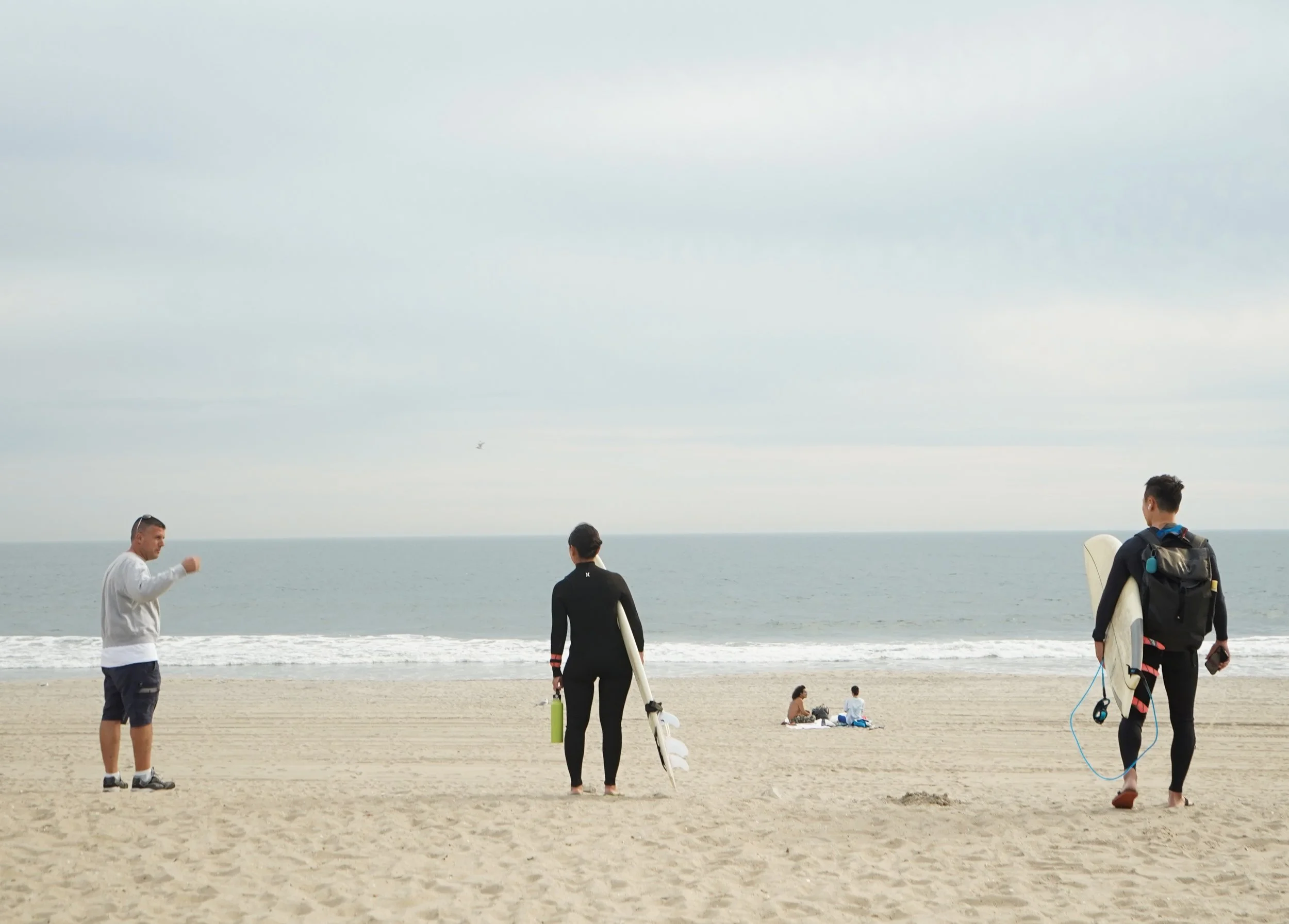Three people standing on a sandy beach, two with surfboards, with the ocean and cloudy sky in the background. Two others are sitting on the sand and the sea is in the distance.