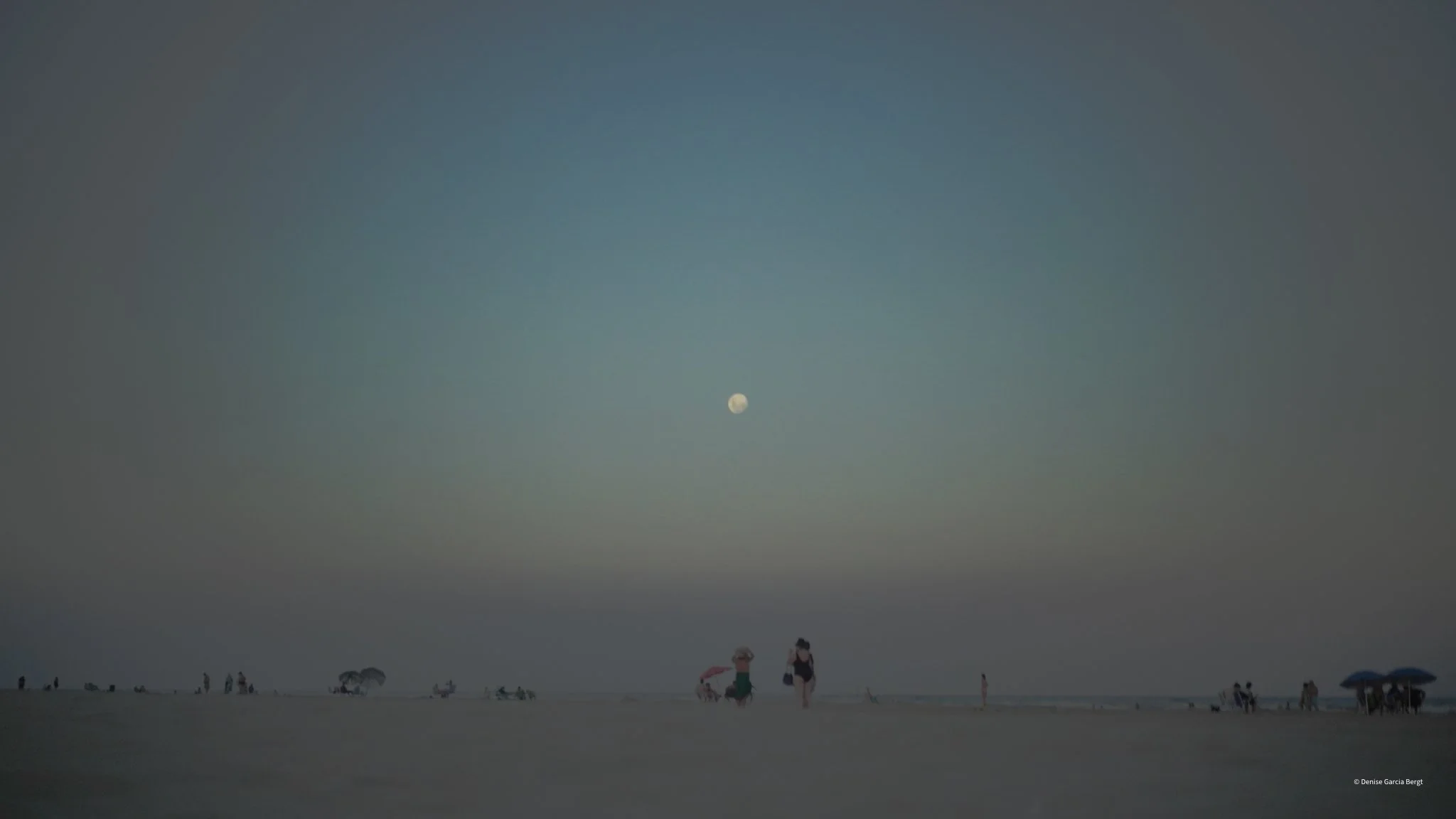 A beach scene at dusk with a clear sky, a visible moon, and a few people relaxing under umbrellas and walking along the shore.