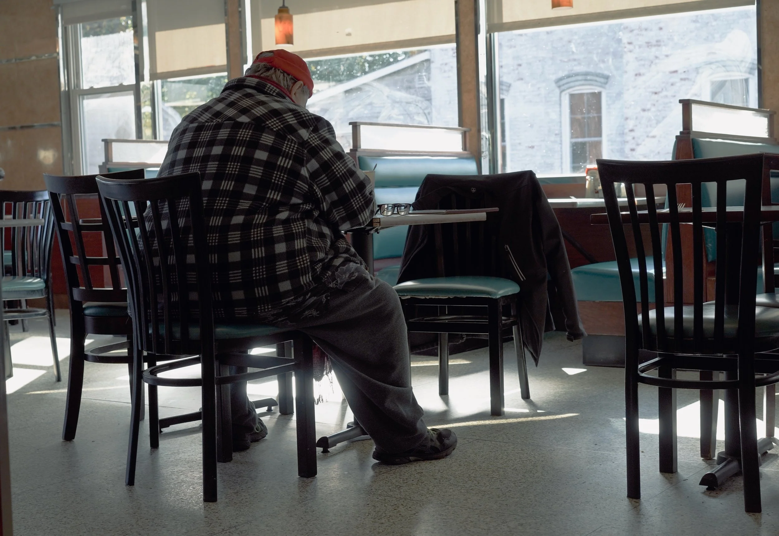 An elderly man in a red cap and checkered jacket sits alone at a table in a restaurant, looking down, with sunglasses resting on the table.