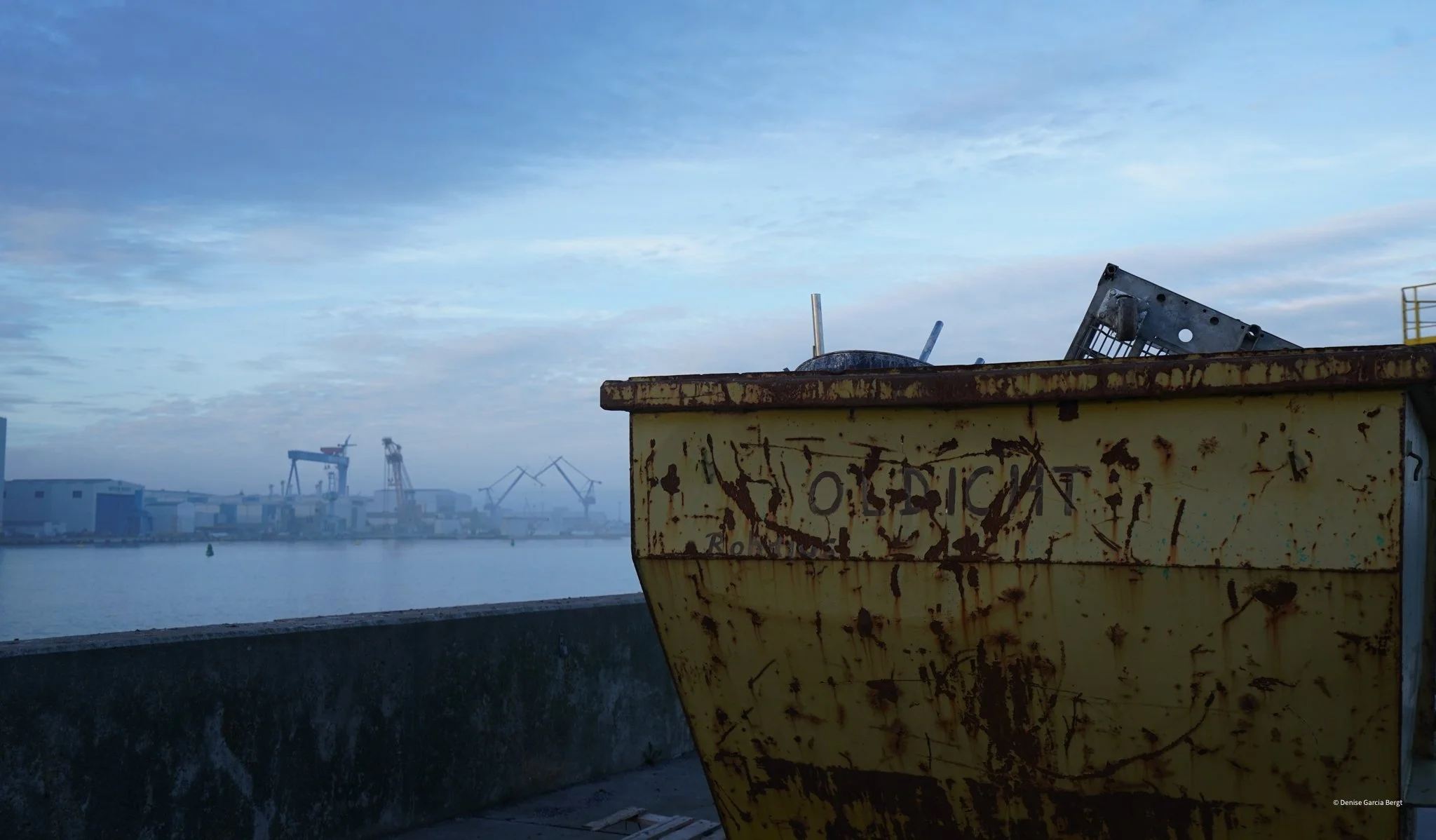 Close-up of a rusted yellow dumpster with discarded metal objects on top, with a harbor and industrial cranes in the background under a cloudy sky.