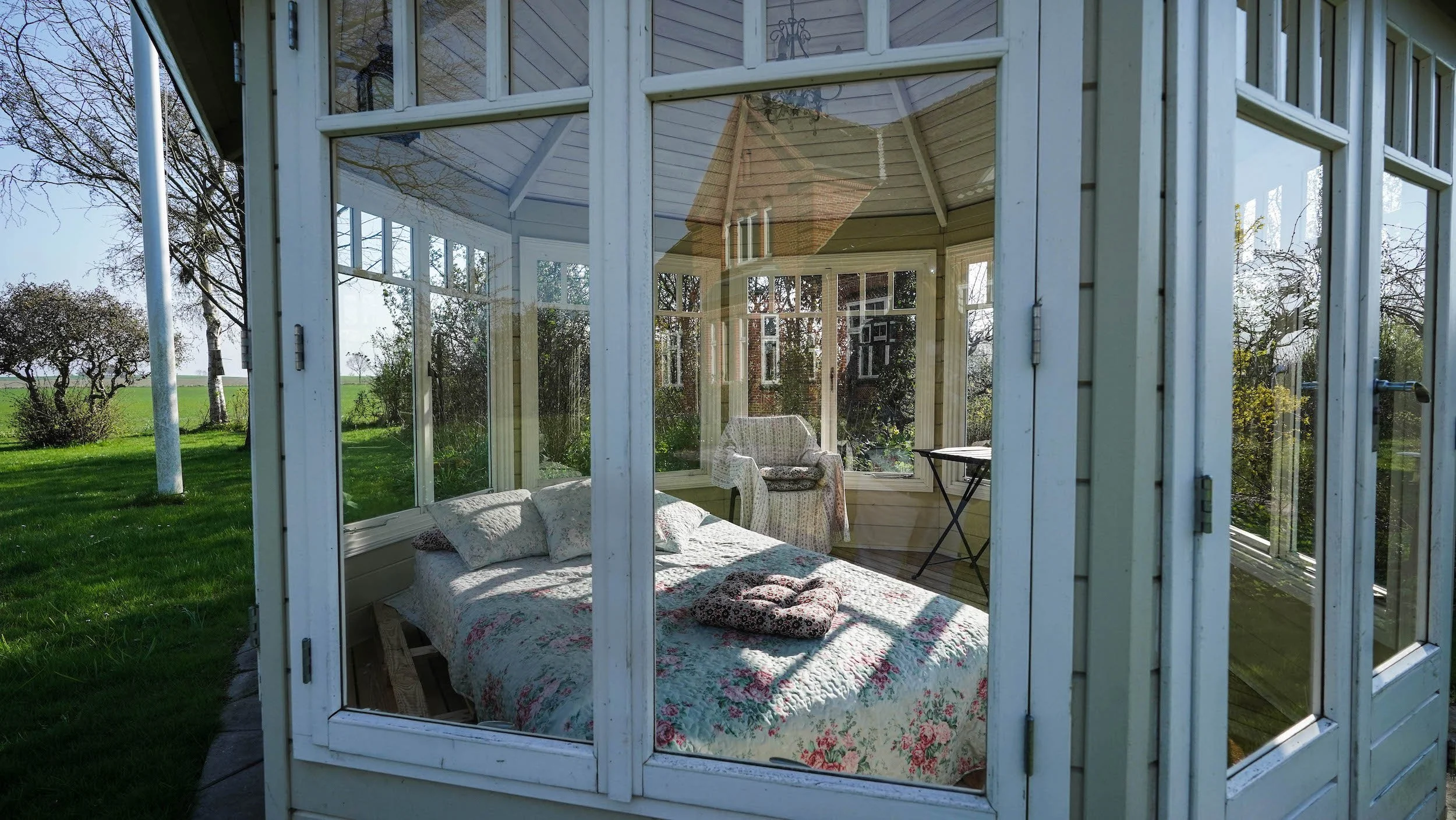 Sunlit wooden sunroom with glass windows and a bed with floral quilt, an armchair, a small table, and a scenic view of trees and green grass outside.
