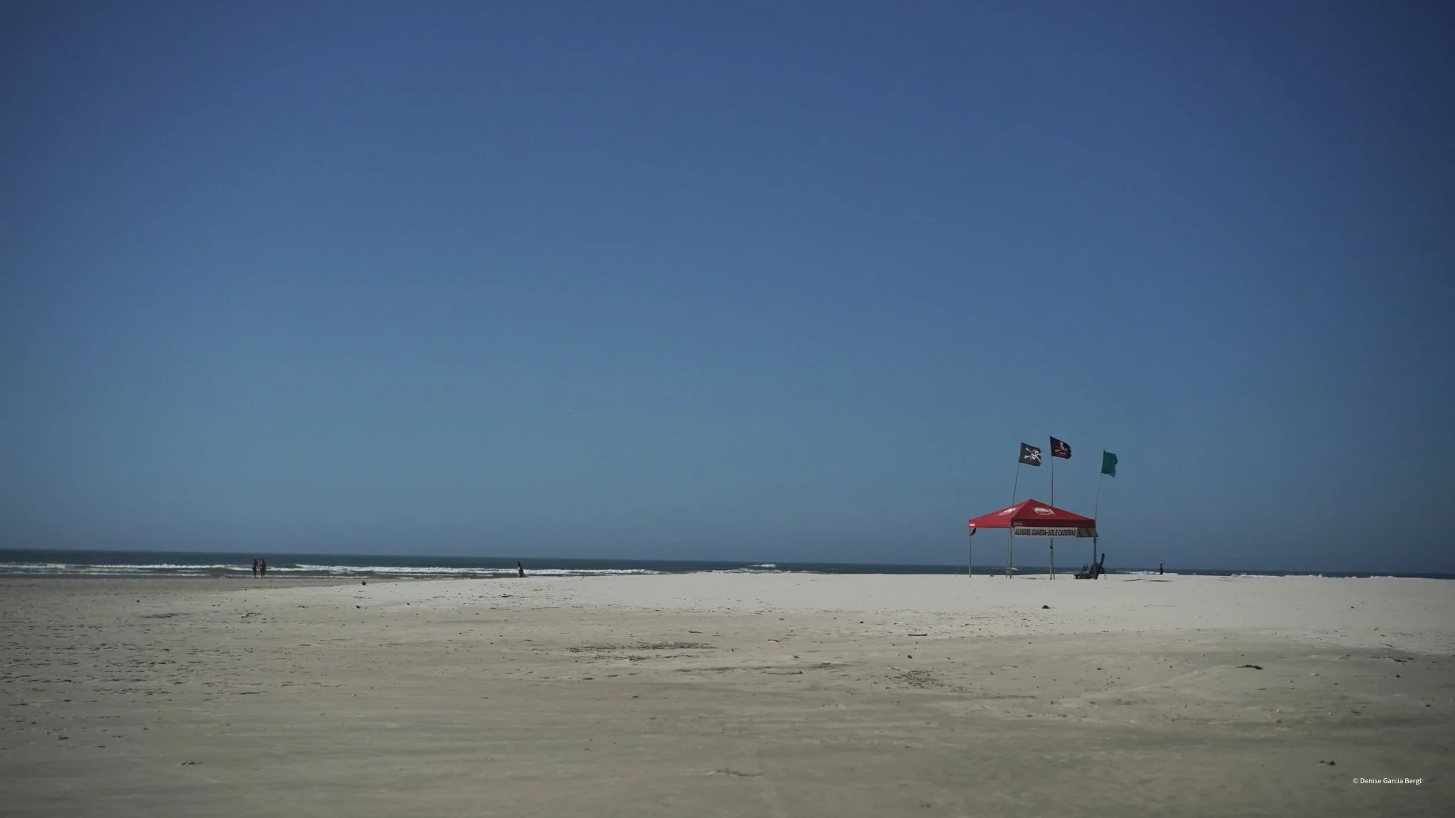 Empty beach with a red canopy and flags, few people walking along the shoreline under a clear blue sky.