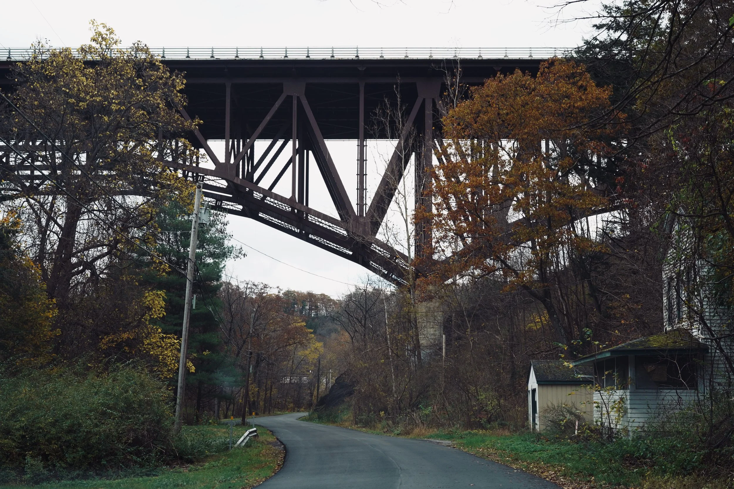 A winding road under a large metal bridge with autumn trees and small houses on the side.