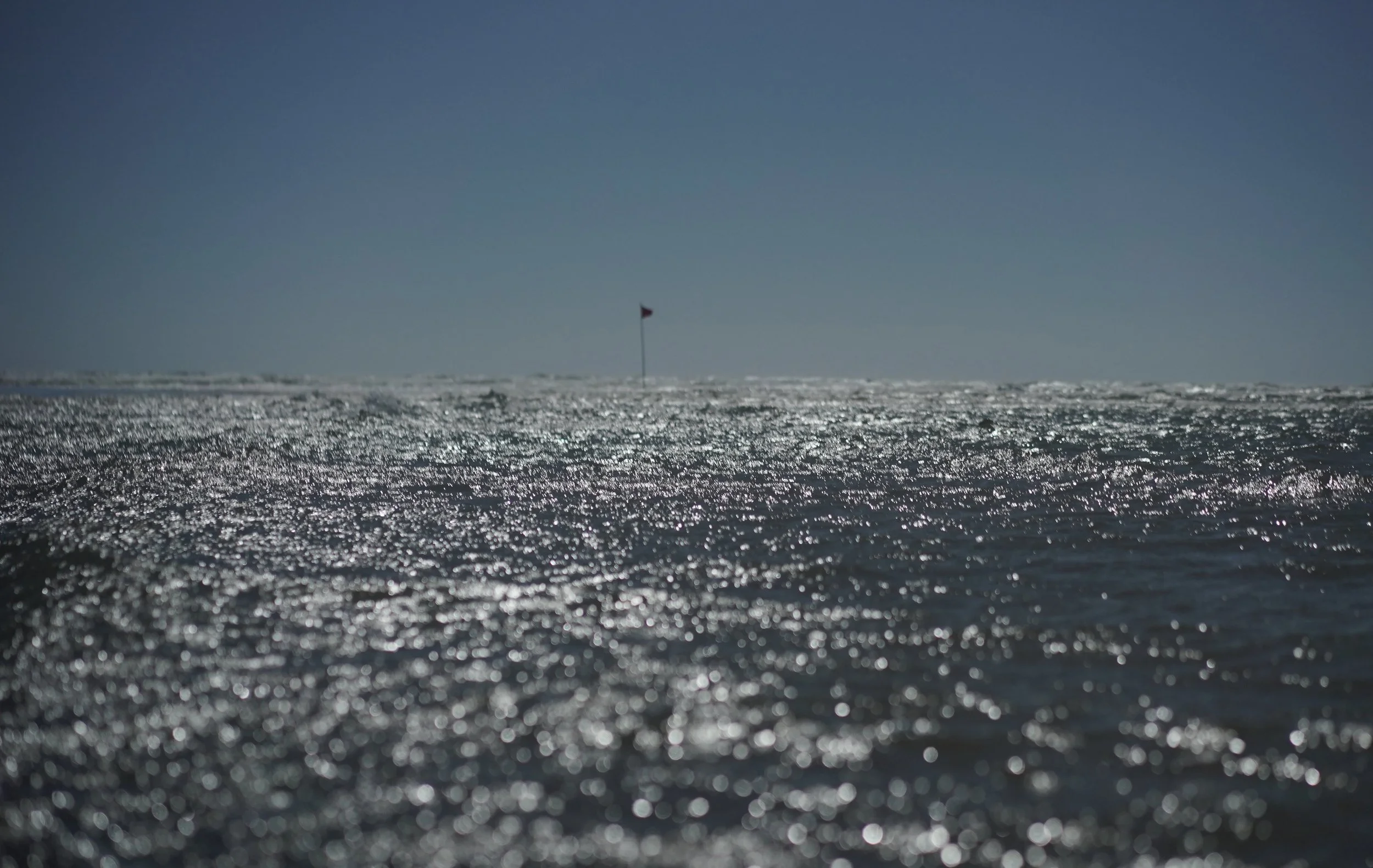 The image shows a vast ocean with sunlight shimmering on the water surface, and a small flag in the distance on the horizon under a clear sky.