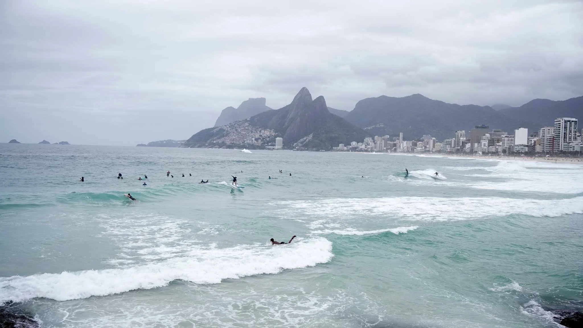 Overcast beach scene in Rio de Janeiro with surfers in the water and city buildings along the shore, mountains in the background.