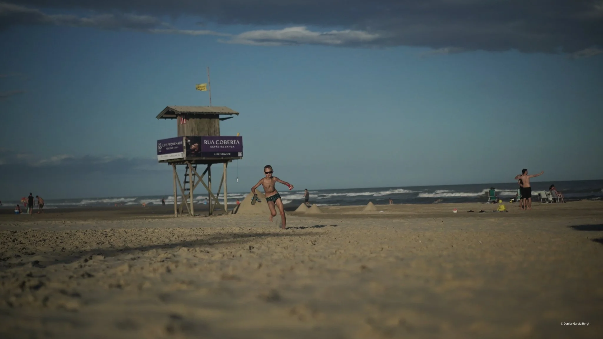A person running on the sandy beach near the ocean, with a lifeguard tower in the background and a few other beachgoers in the distance.