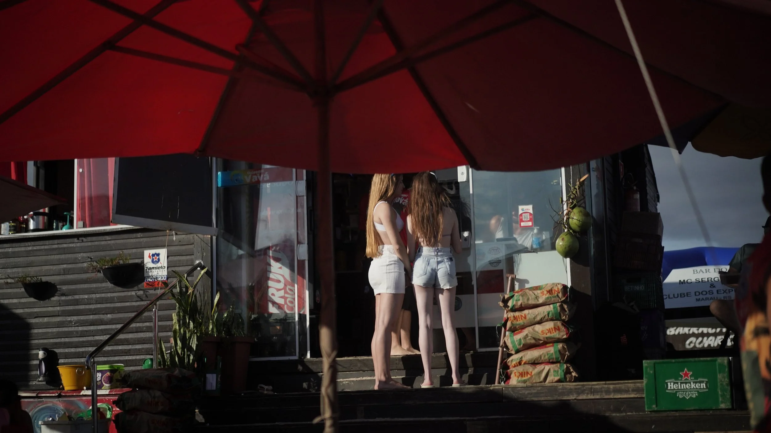 Two women with long hair wearing denim shorts are standing inside a small store or kiosk, visible through a glass door, under an umbrella.