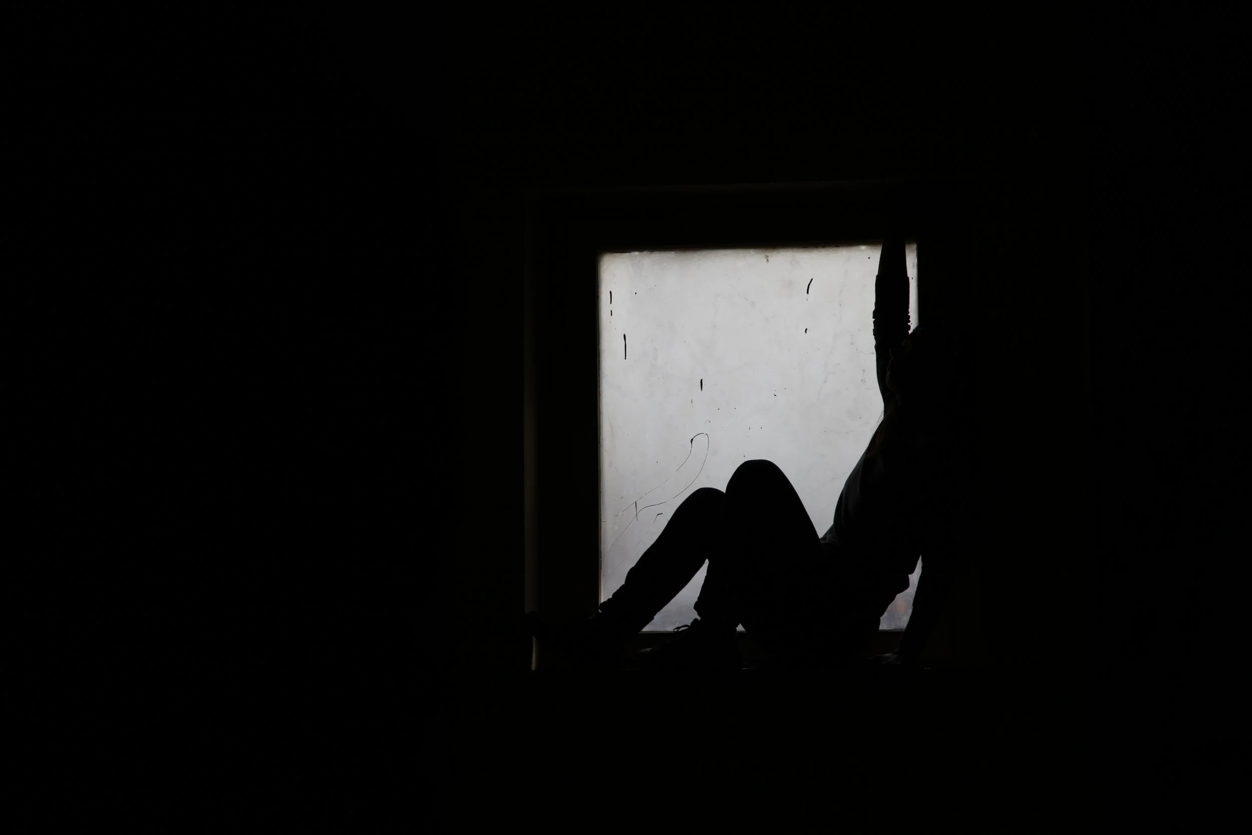 Silhouette of a person sitting on a window sill with their knees up, facing out, in a dark room with light coming through the window.