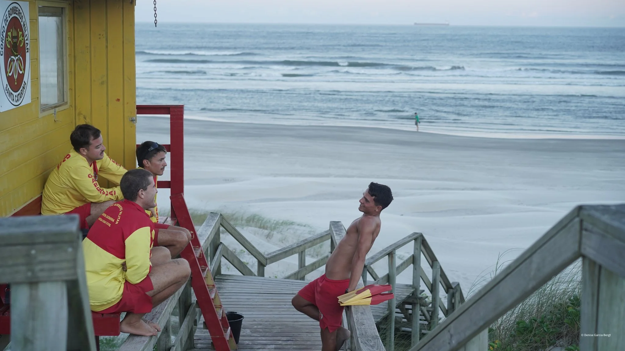 Four lifeguards in yellow and red uniforms sitting and standing on a wooden staircase at the beach, engaging in conversation. One lifeguard is shirtless, holding red and yellow floatation devices, leaning on the railing. The sandy beach and ocean wav