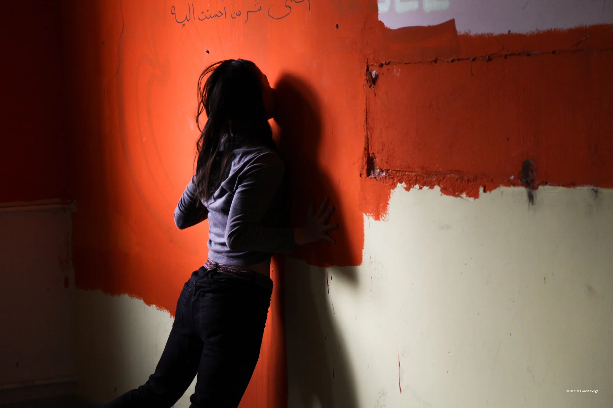 A woman with dark hair is standing against a wall, painting it orange. Her face is not visible, and she is wearing a gray long-sleeve shirt and black pants. The wall has a section of orange paint partially applied over a cream-colored surface.