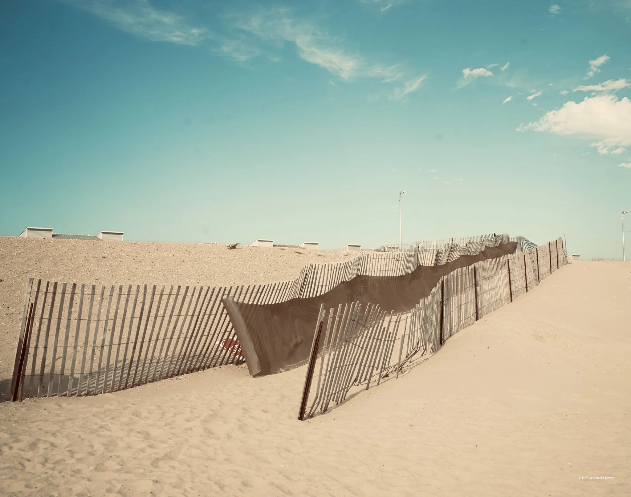 A fence on a sandy beach that is damaged and falling down, with sand dunes and a clear blue sky in the background.