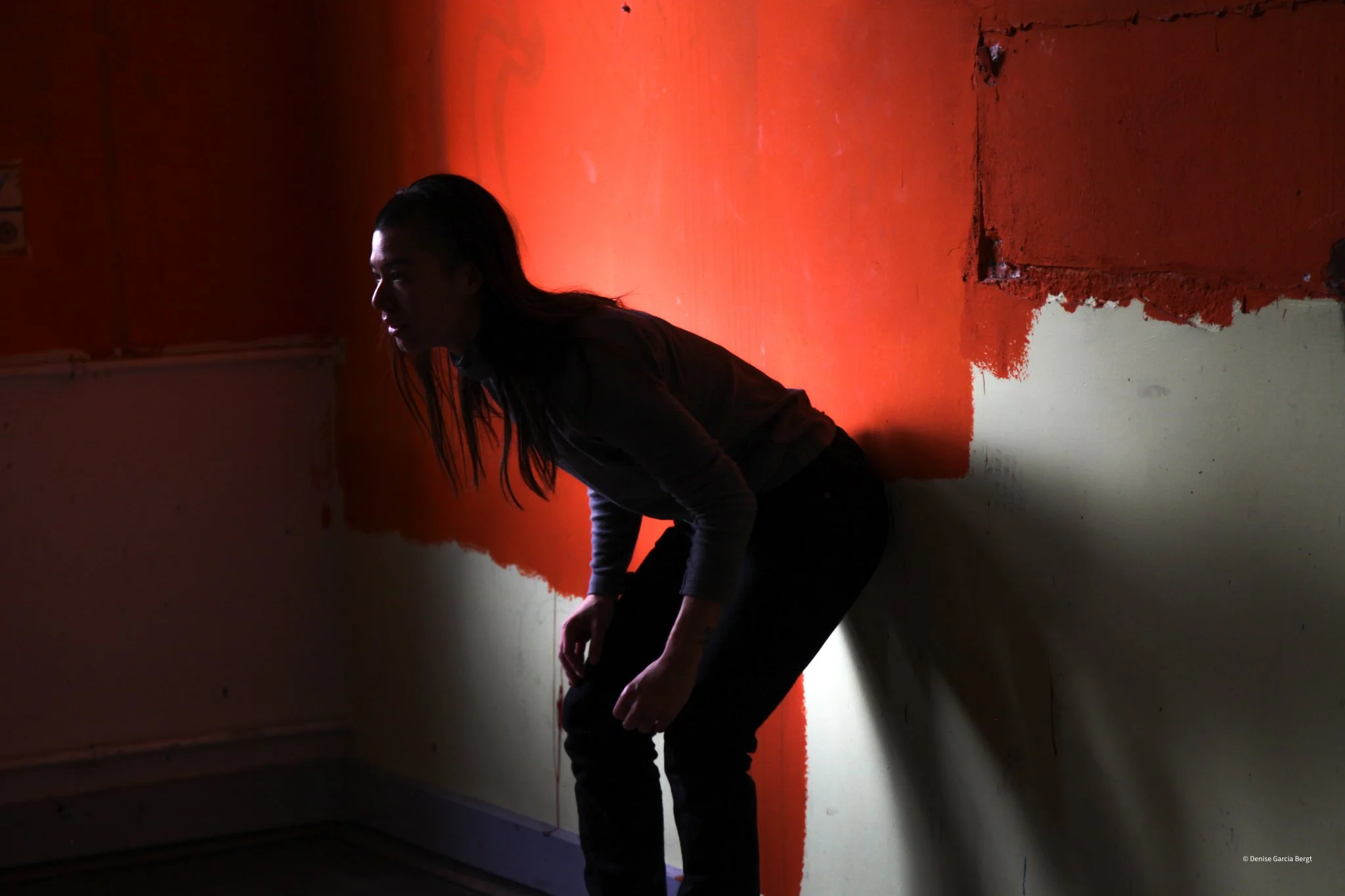 A person with long hair bent over, sitting against a wall with red and beige paint. The room is dimly lit.