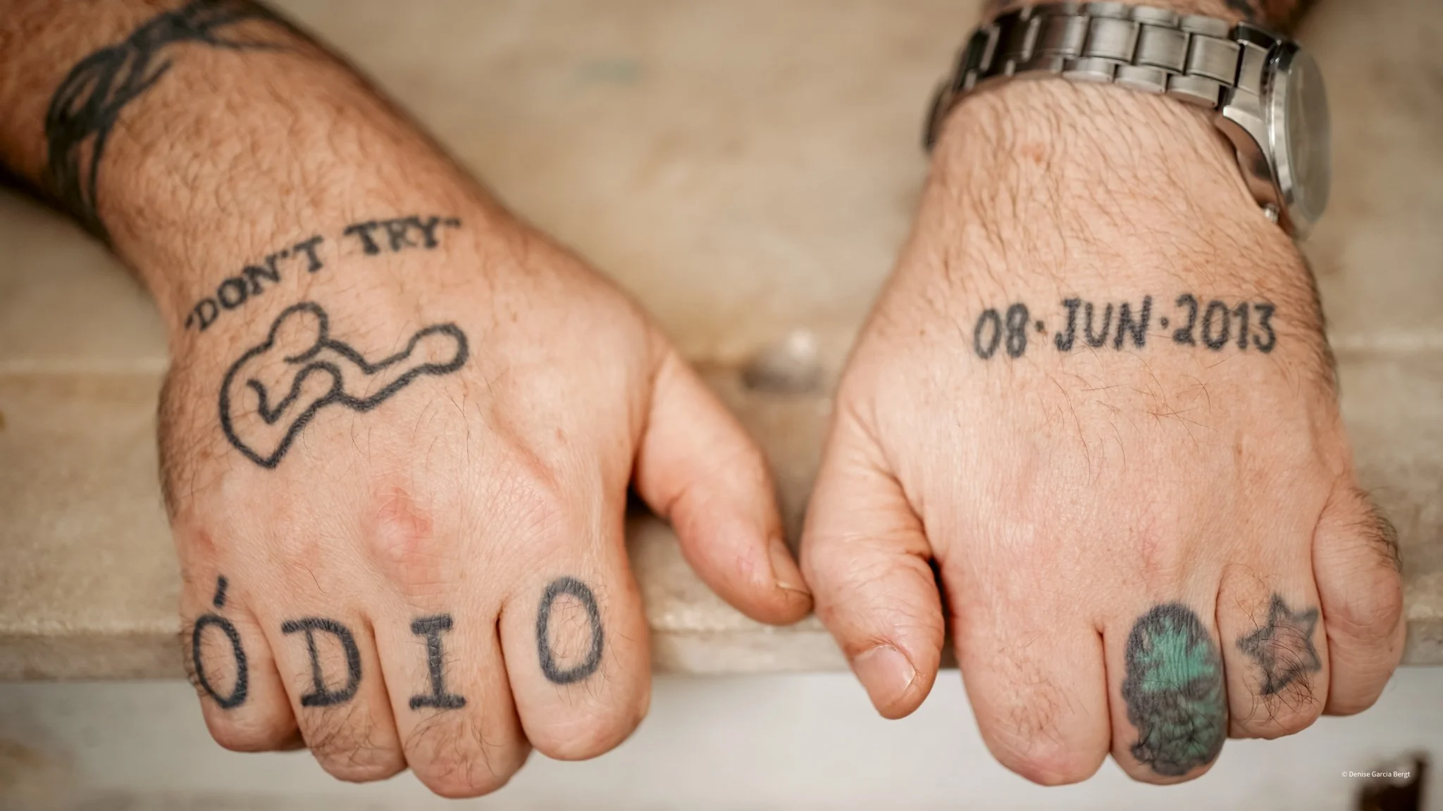 Close-up of tattooed hands with the words 'DON'T TRY', a boxing glove outline, 'ÓDIO', the date '08 JUN 2013', and small star and leaf tattoos.