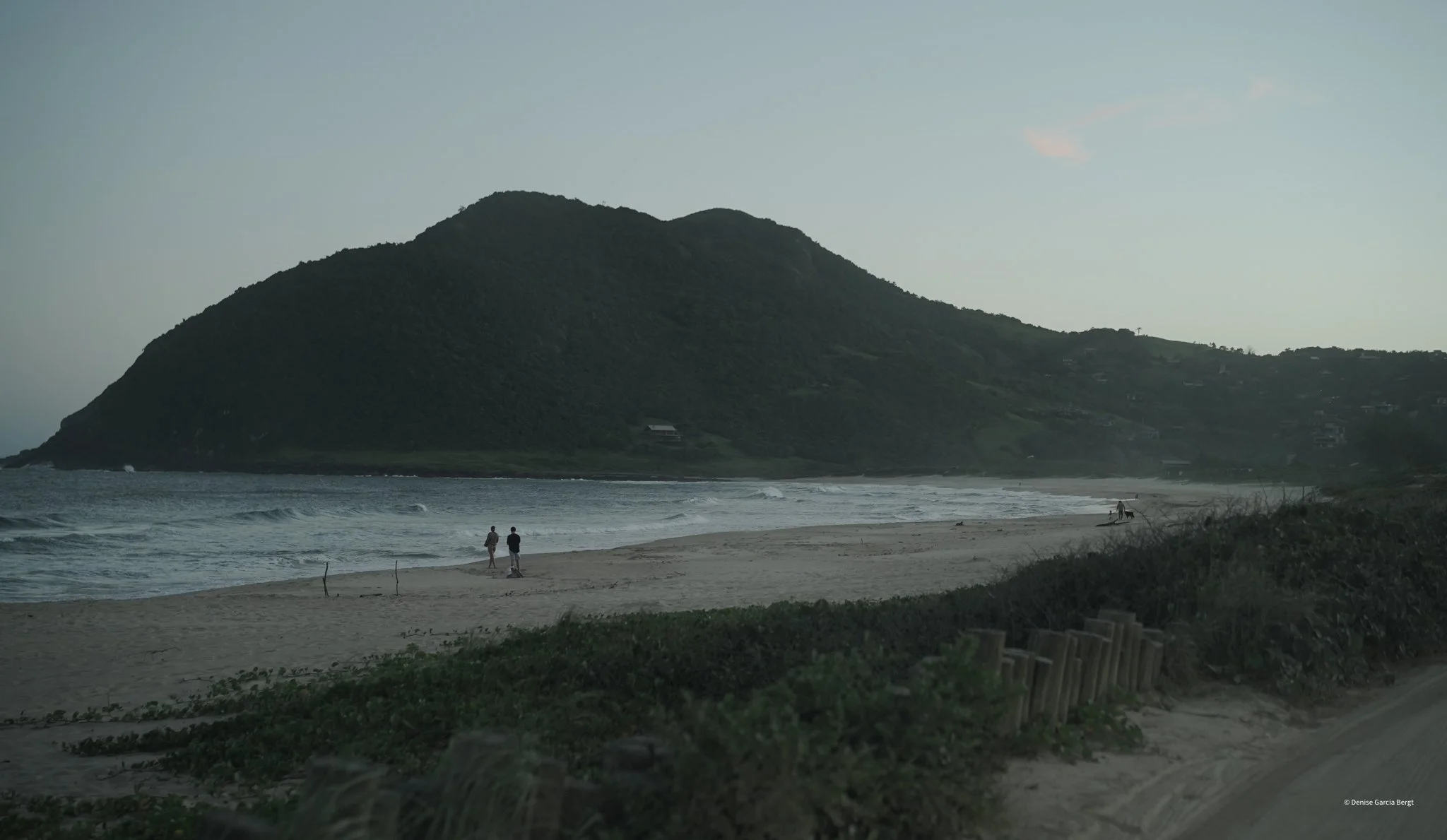 A serene beach scene at dusk with a large green hill or small mountain in the background, gentle waves approaching the shore, and a few people walking near the water.