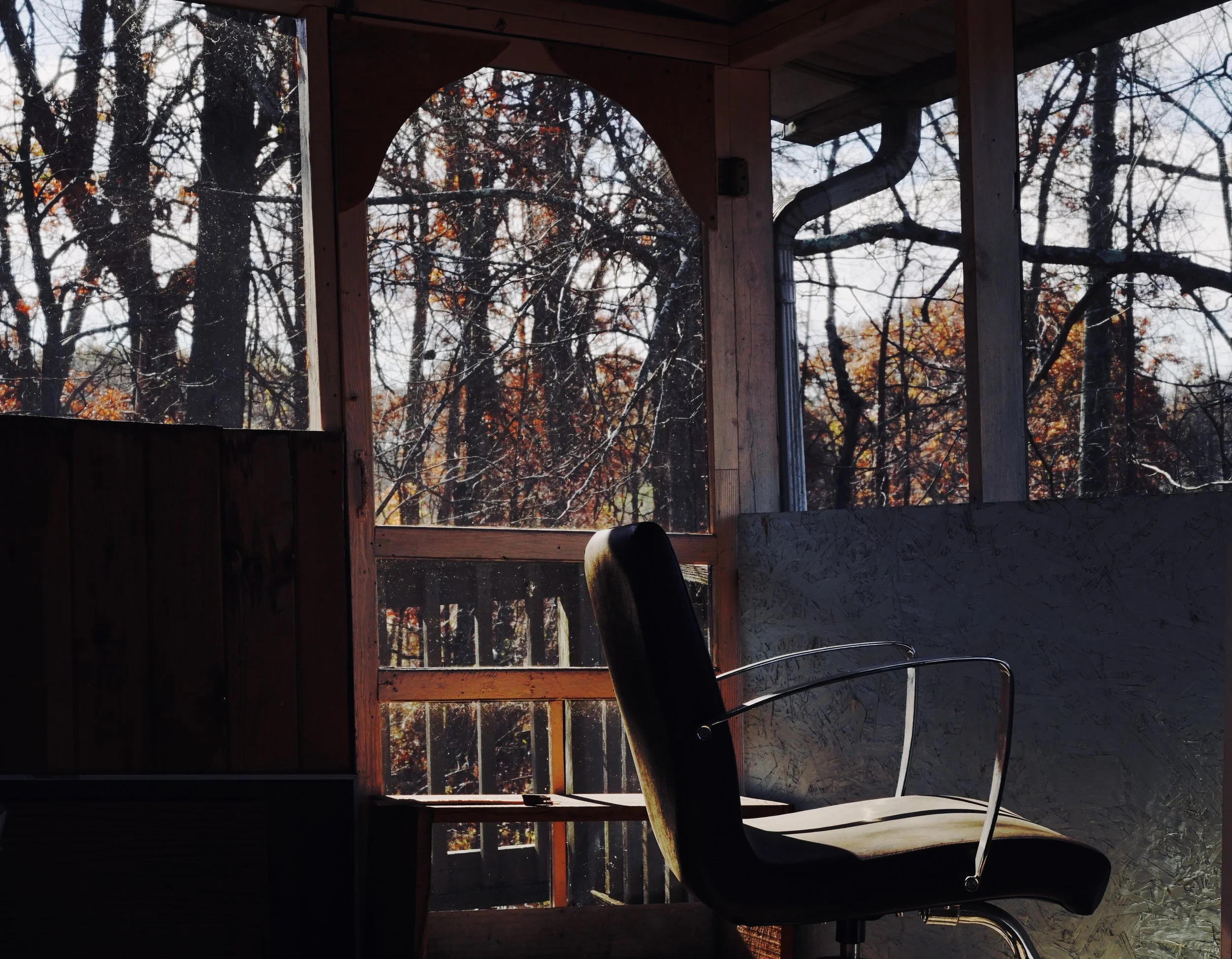 A black office chair with metal armrests in a rustic wooden balcony or porch, with a view of leafless trees and a partly cloudy sky outside.
