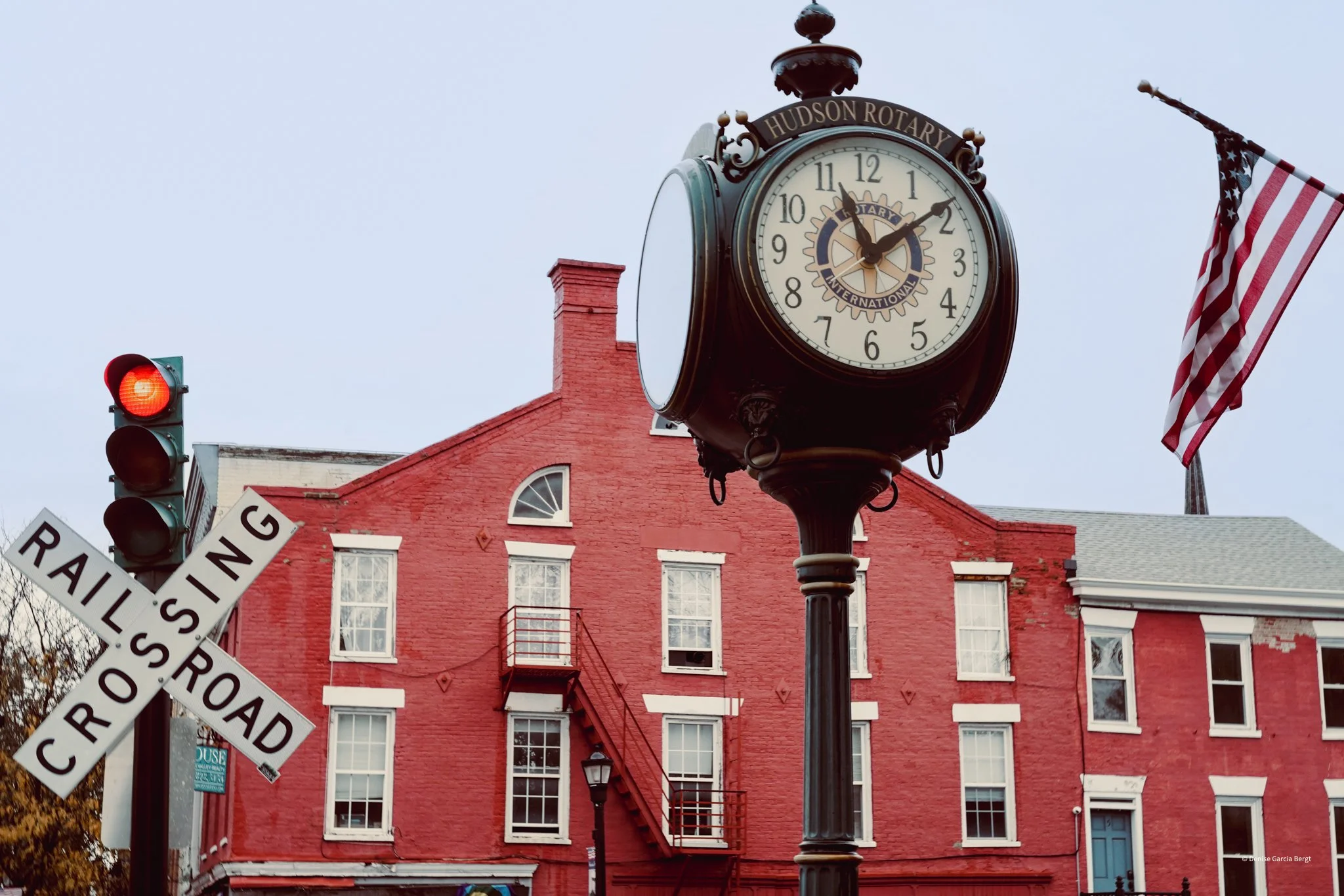 A street scene featuring a large vintage clock on a black post with the words 'Hudson Rotary' on top, an American flag waving in the background, a red brick building with multiple windows, an external fire escape, a red traffic light, and a train cro