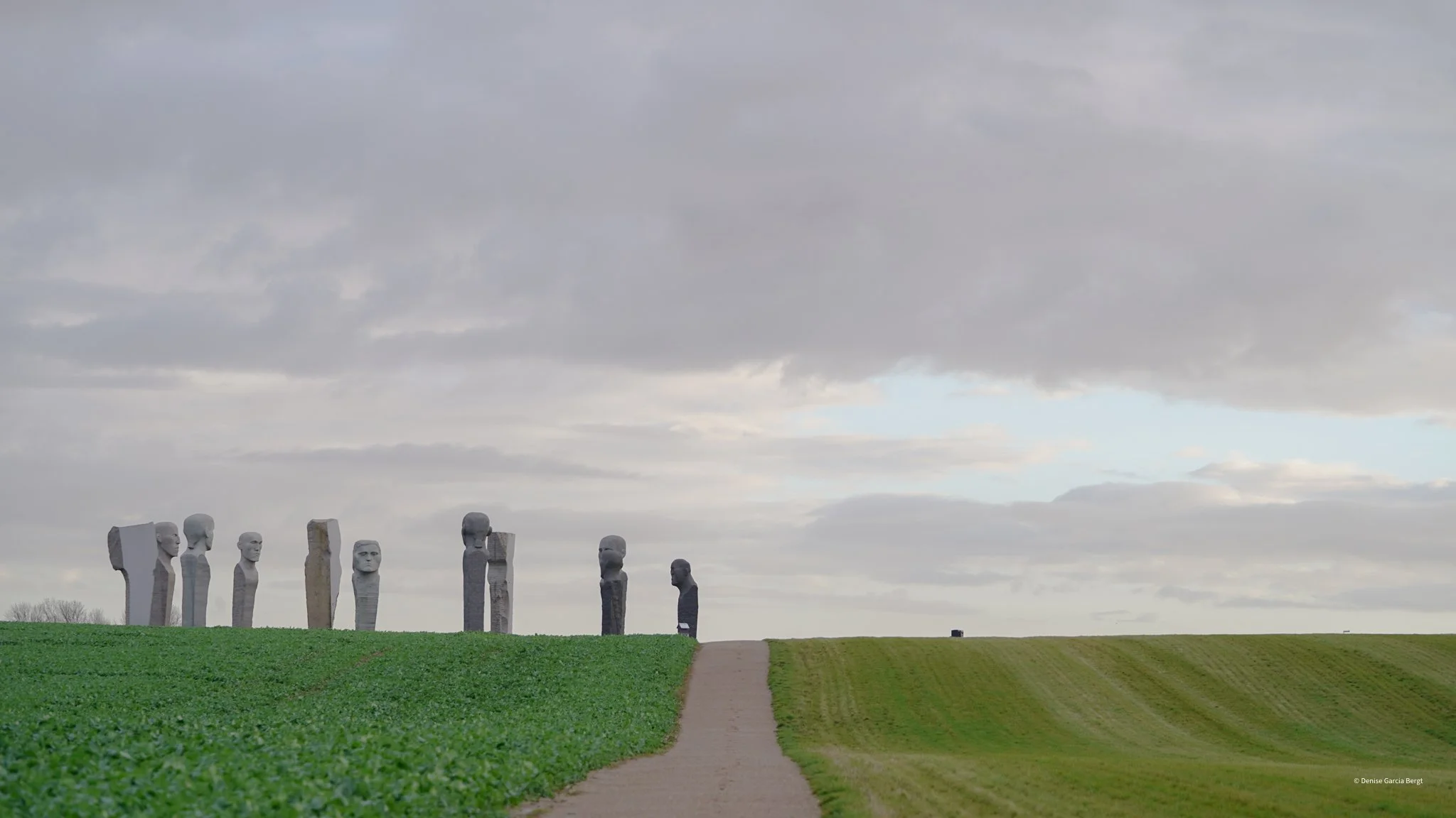 A walkway through green fields leads to a monument of several tall stone statues resembling heads and torsos, with a person walking towards the monument, under a cloudy sky.