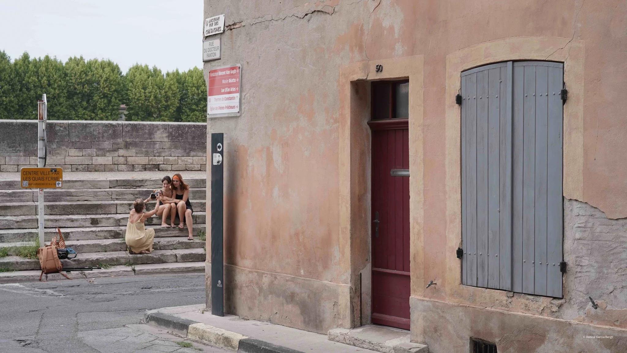 Two women sitting on stone steps with an orange-haired woman taking a photo of them as a young girl kneels in front of her. Bags and a tripod are on the sidewalk nearby. The building has a closed gray wooden shutter and a door with the number 50 on t