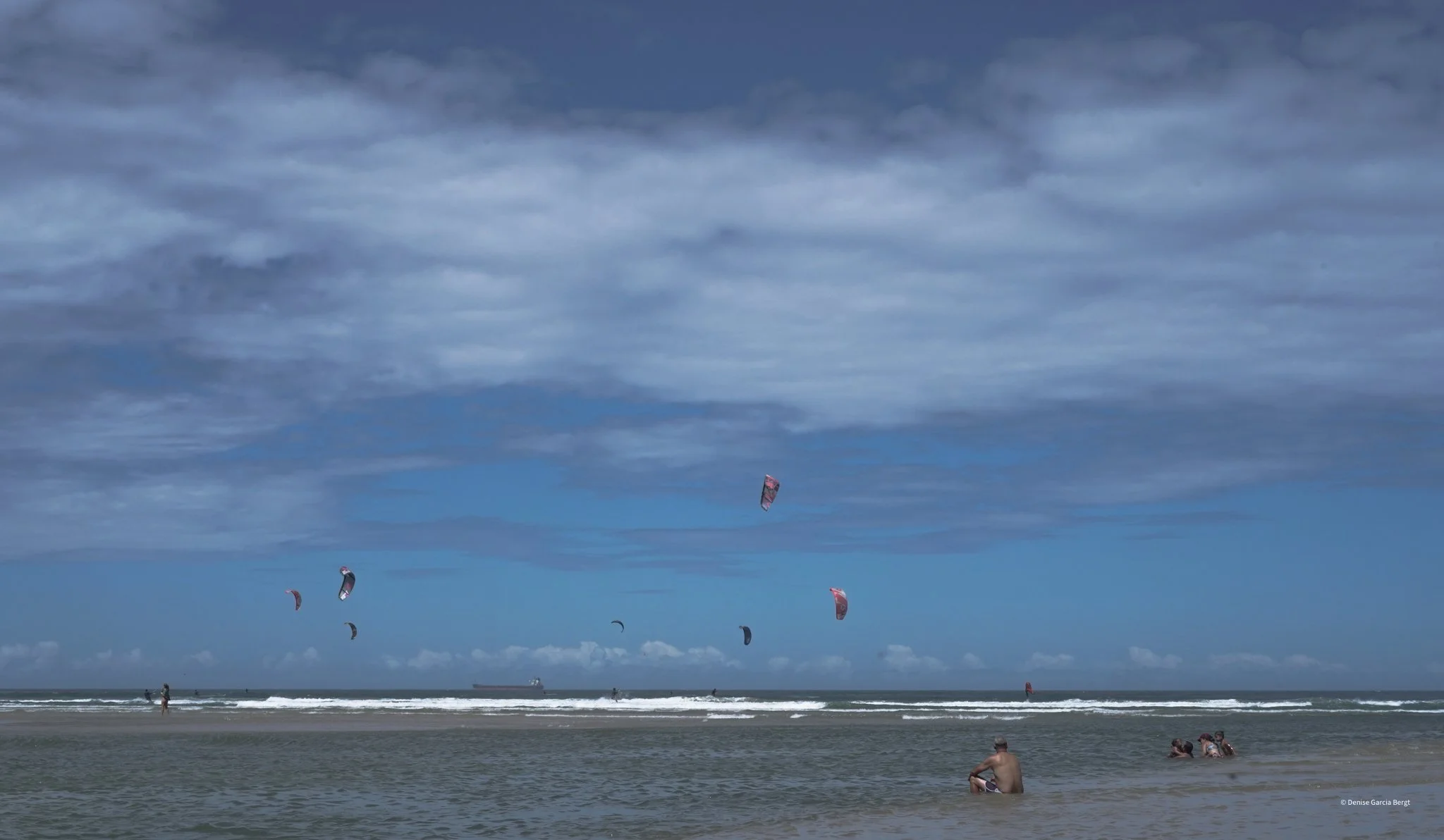 People kite surfing and relaxing on the beach with a ship in the distance under a cloudy sky.