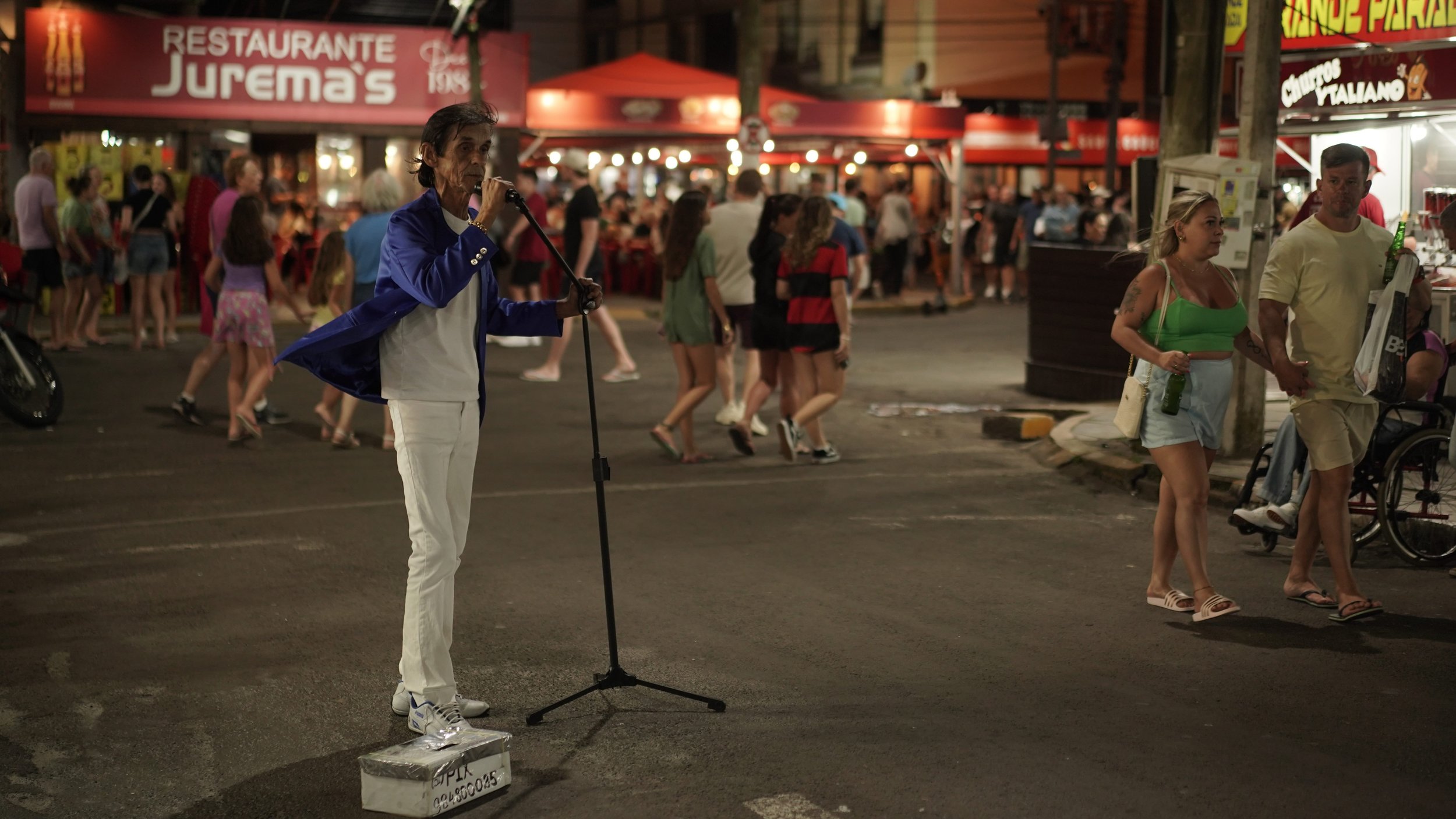 A street scene at night with a man in a blue blazer, white shirt, and white pants singing into a microphone on a stand, standing on a small platform. In the background, there are many people walking and socializing near food stalls and a restaurant w