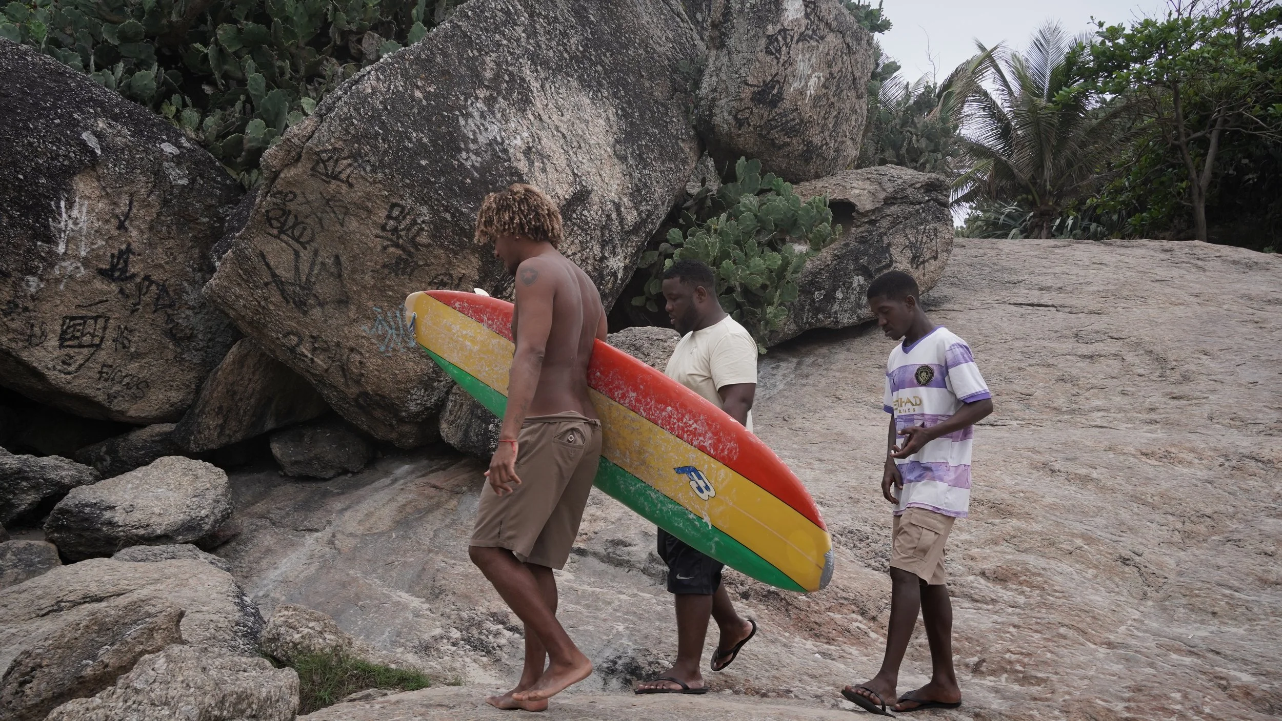 Three young men walk along rocky terrain, one holding a colorful surfboard, with large boulders and green trees in the background.