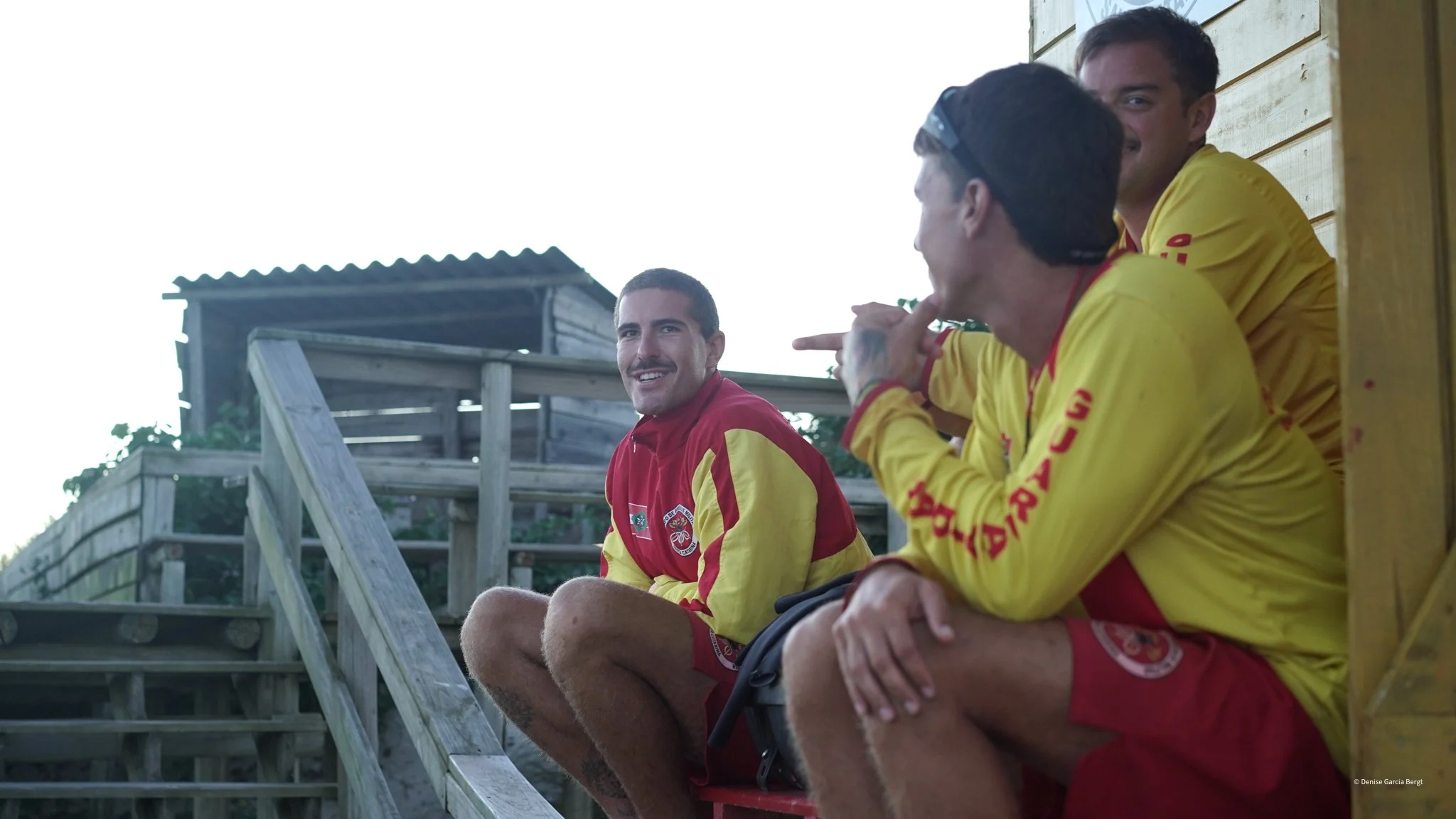 Three young men in red and yellow sports jackets sitting on wooden stairs outside, engaged in conversation and smiling.