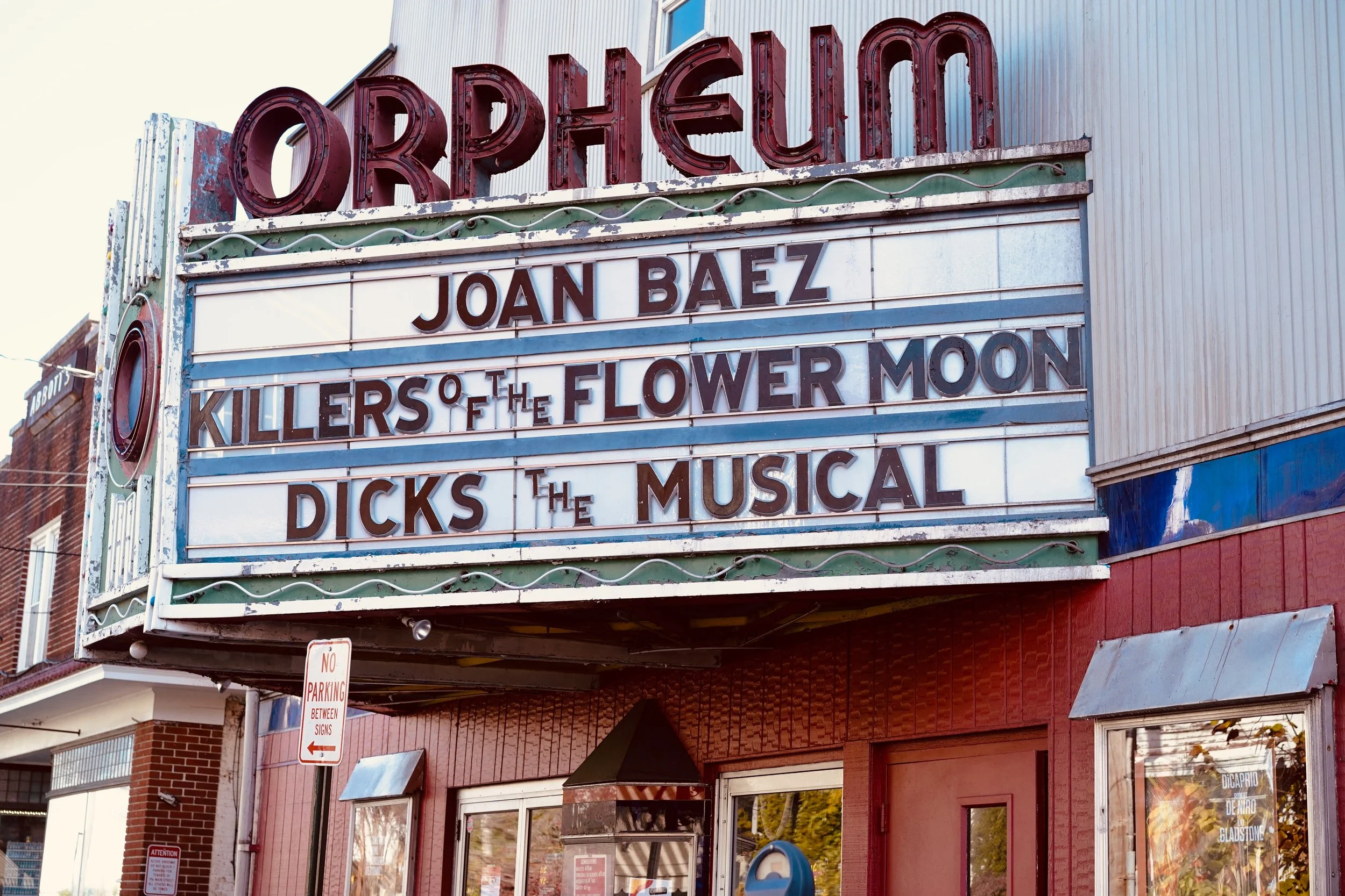 An old theater marquee with a sign featuring the names of acts: Joan Baez, Killers of the Flower Moon, Dicks the Musical, and others, on a building with a brick and metal facade.