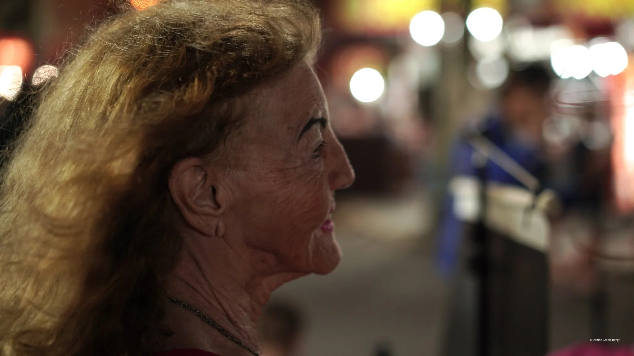 Profile of an elderly woman with long, wavy blonde hair, smiling indoors at night with blurred lights and people in the background.