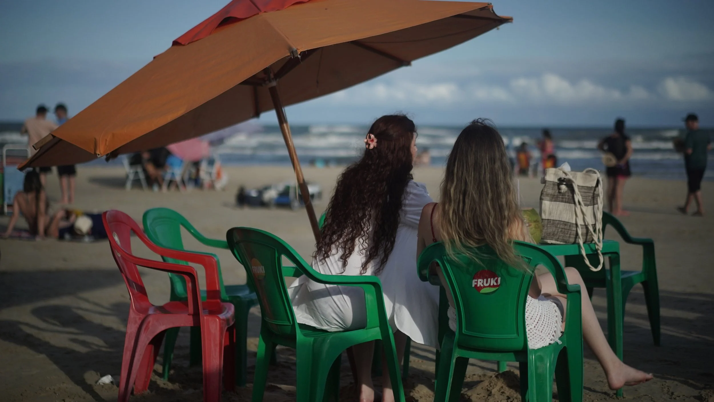 Two women sitting on green and red plastic chairs under a large beach umbrella on the sandy beach, with the ocean in the background.
