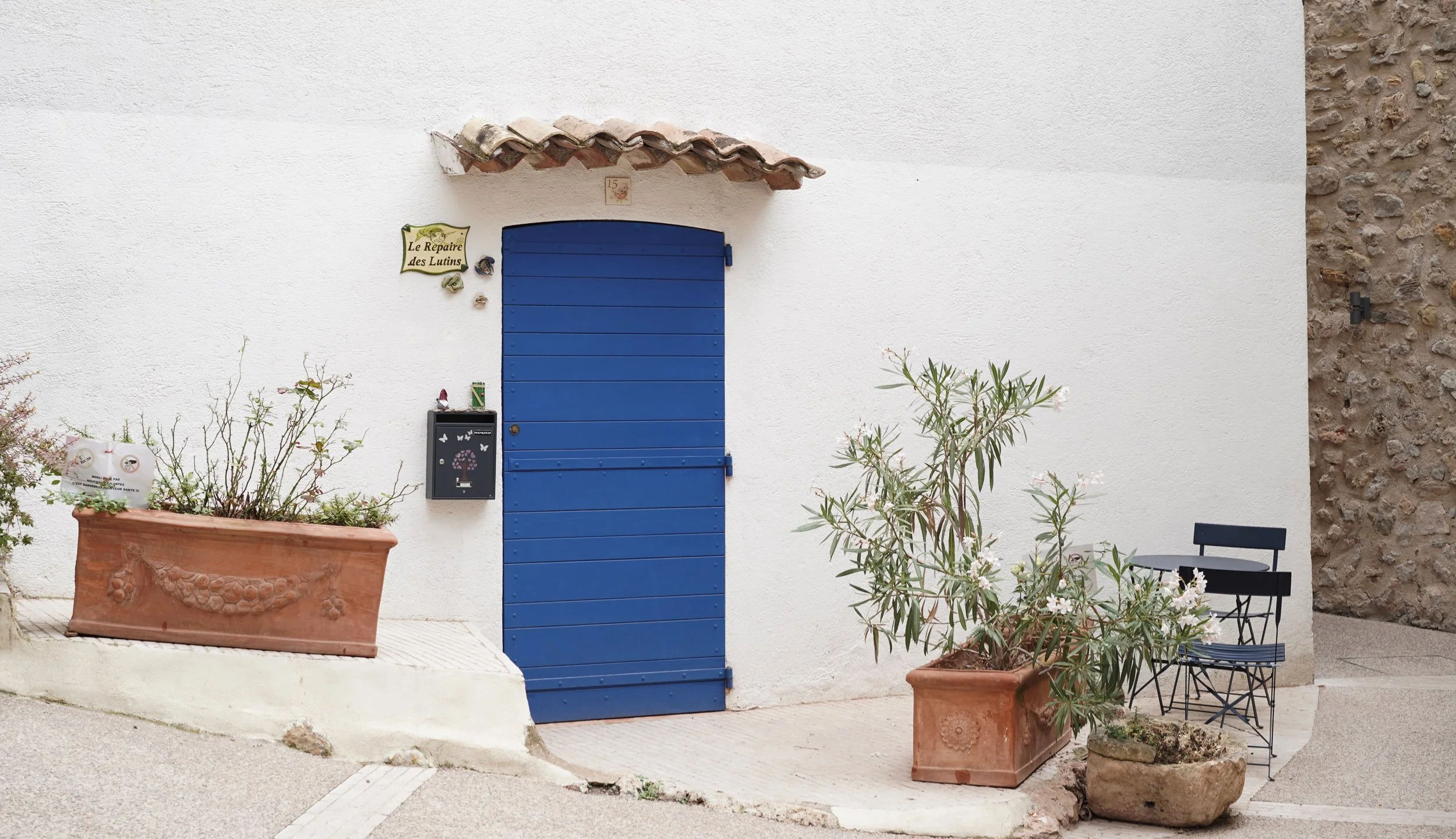 White wall with a blue door, two large terracotta planters with green plants, a black chair and small table, and a small sign reading "Le Repaire des Lutins".
