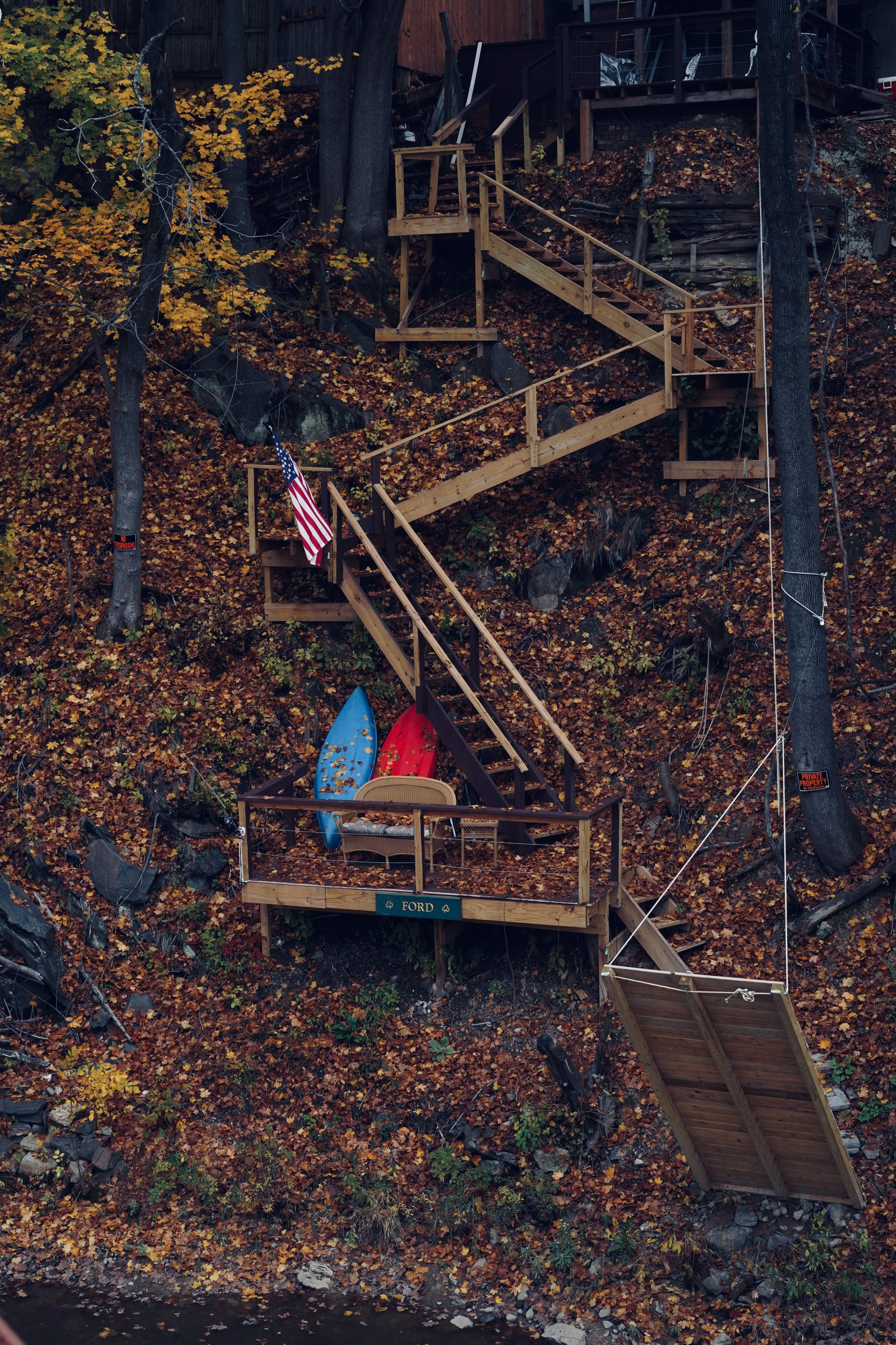 A wooden staircase ascending a steep, leaf-covered hillside, with a dock at the bottom holding two paddles and a chair. An American flag is attached to the staircase railing near the top.