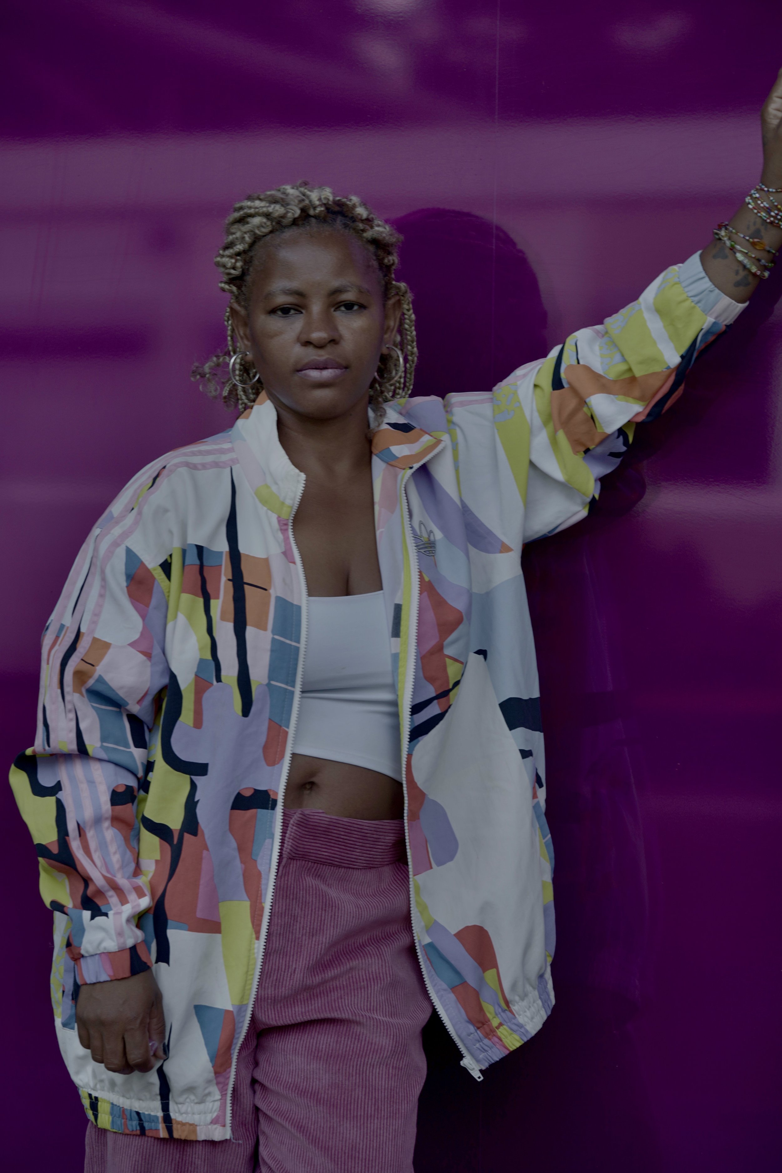 Woman with blonde, styled hair and hoop earrings, wearing a colorful patterned jacket over a white crop top, pink corduroy pants, standing against a purple wall.