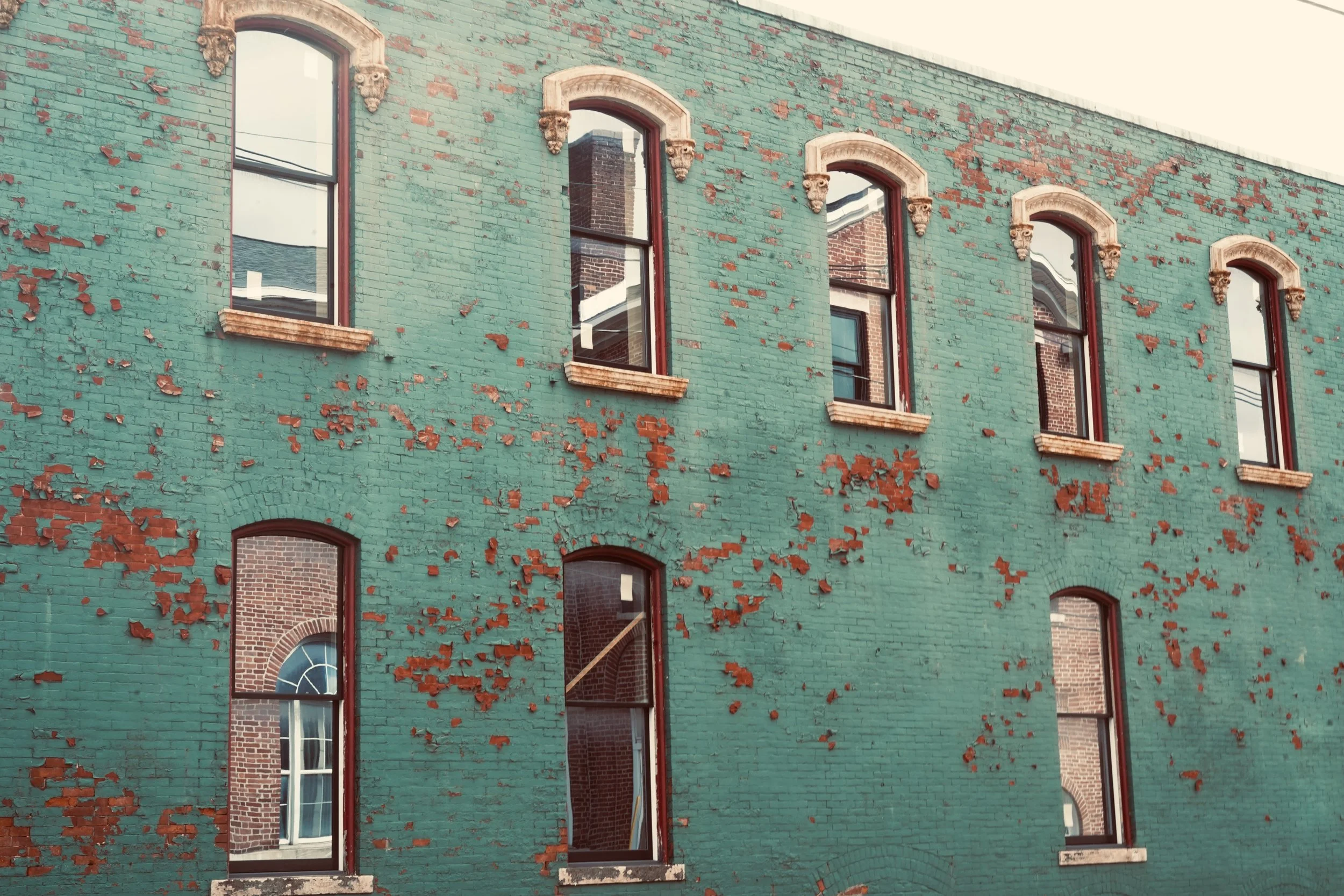 A brick building with a green painted exterior, showing signs of peeling paint and weathering. It has six windows with decorative brickwork and carved stone accents above each window.