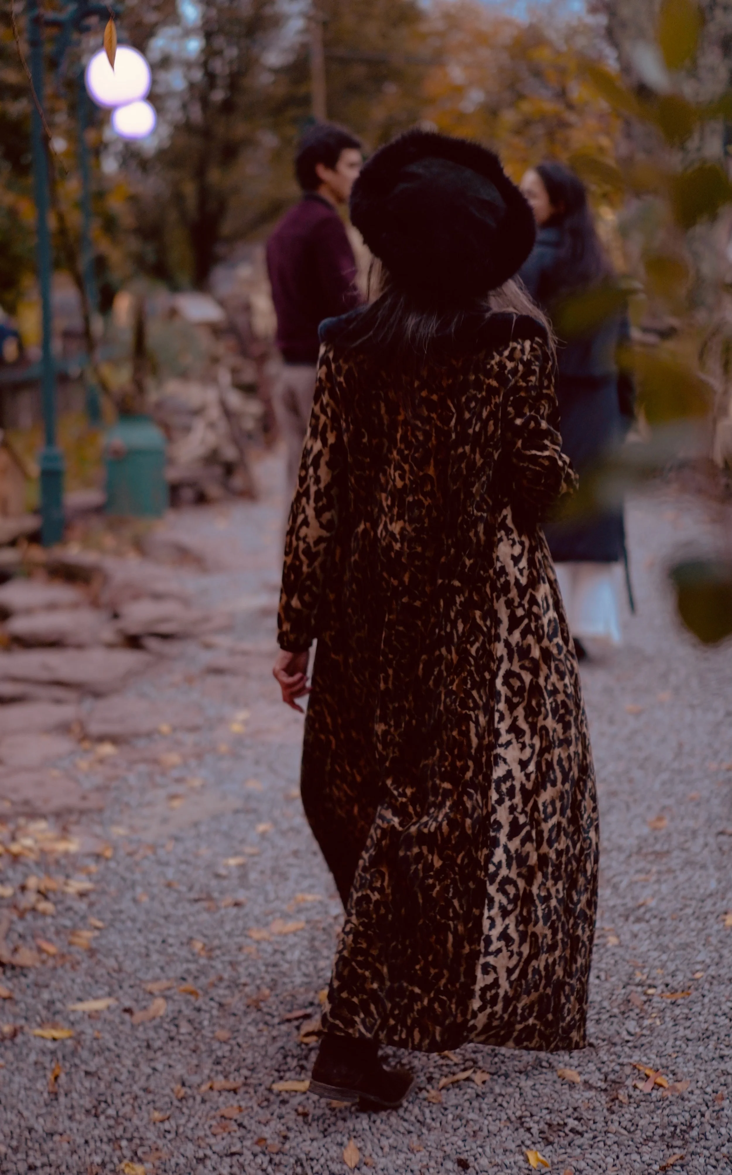 A woman wearing a leopard print coat and a black hat walking on a gravel path during fall, with two people in the background talking.