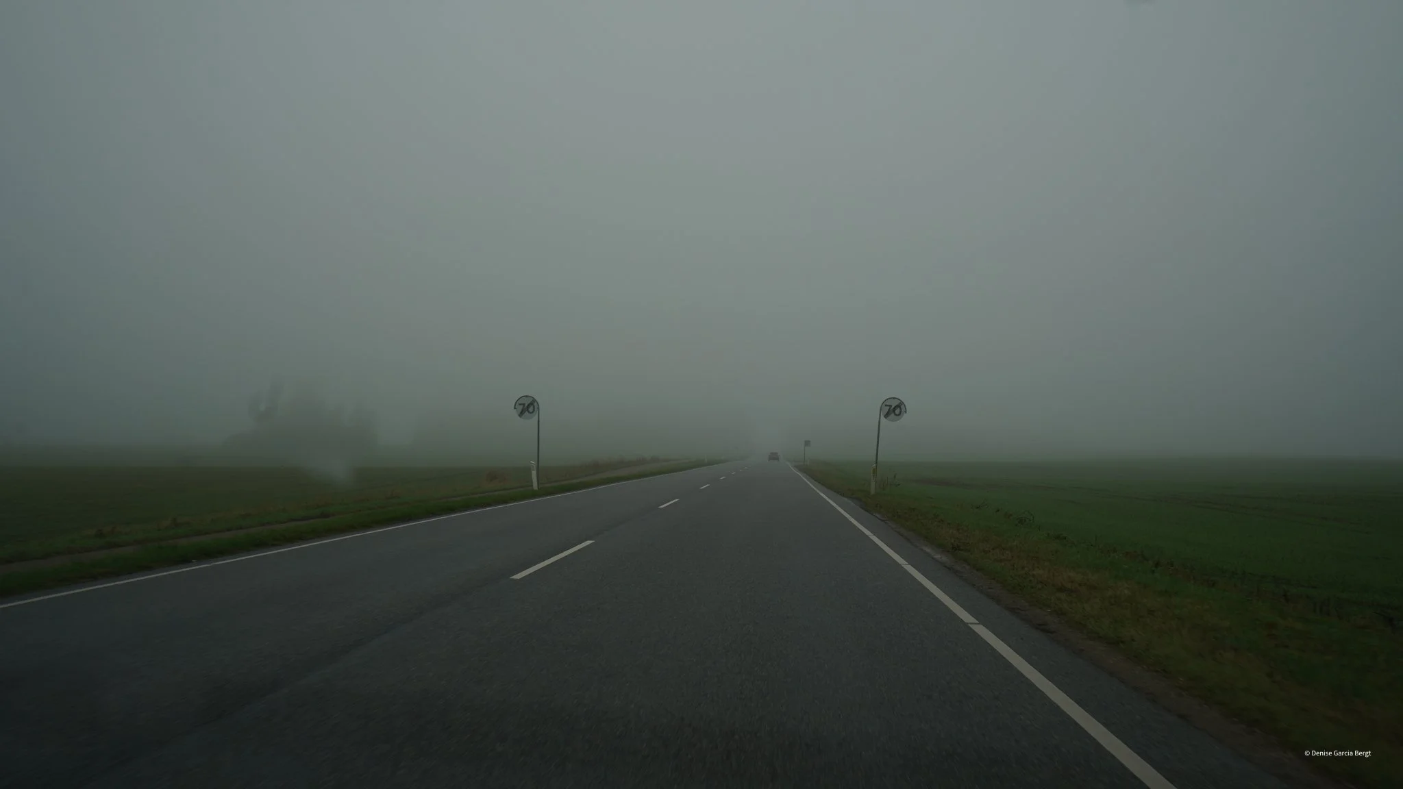 A foggy road with two speed limit signs showing 70 km/h, and visibility is low due to dense fog. The road runs through a flat green field on both sides.