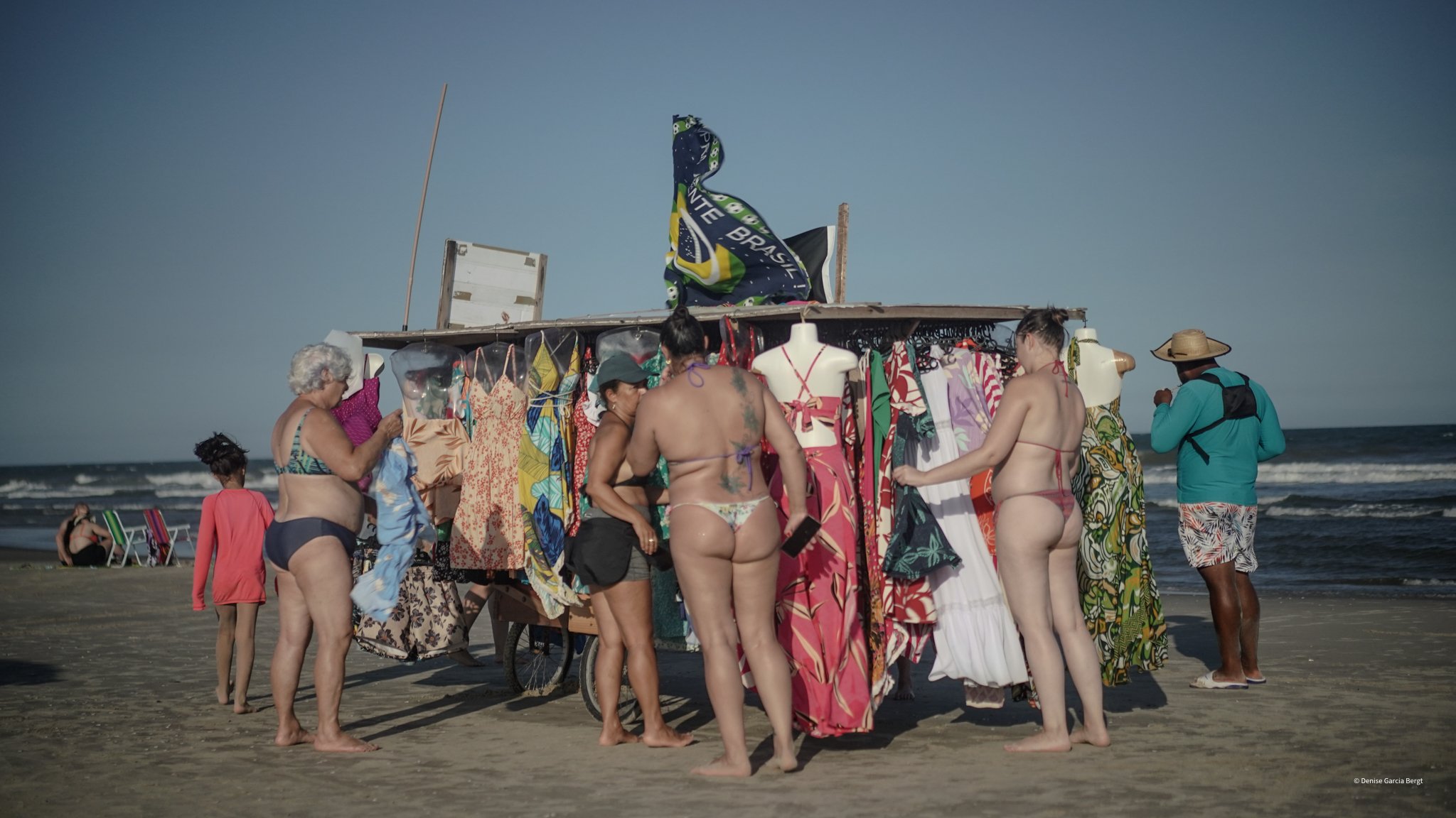 People shopping at a beachside market with colorful clothing on mannequins, on a sandy beach with the ocean in the background. Several women in swimsuits, some with tattoos, and a man in a blue shirt and hat are browsing the merchandise.
