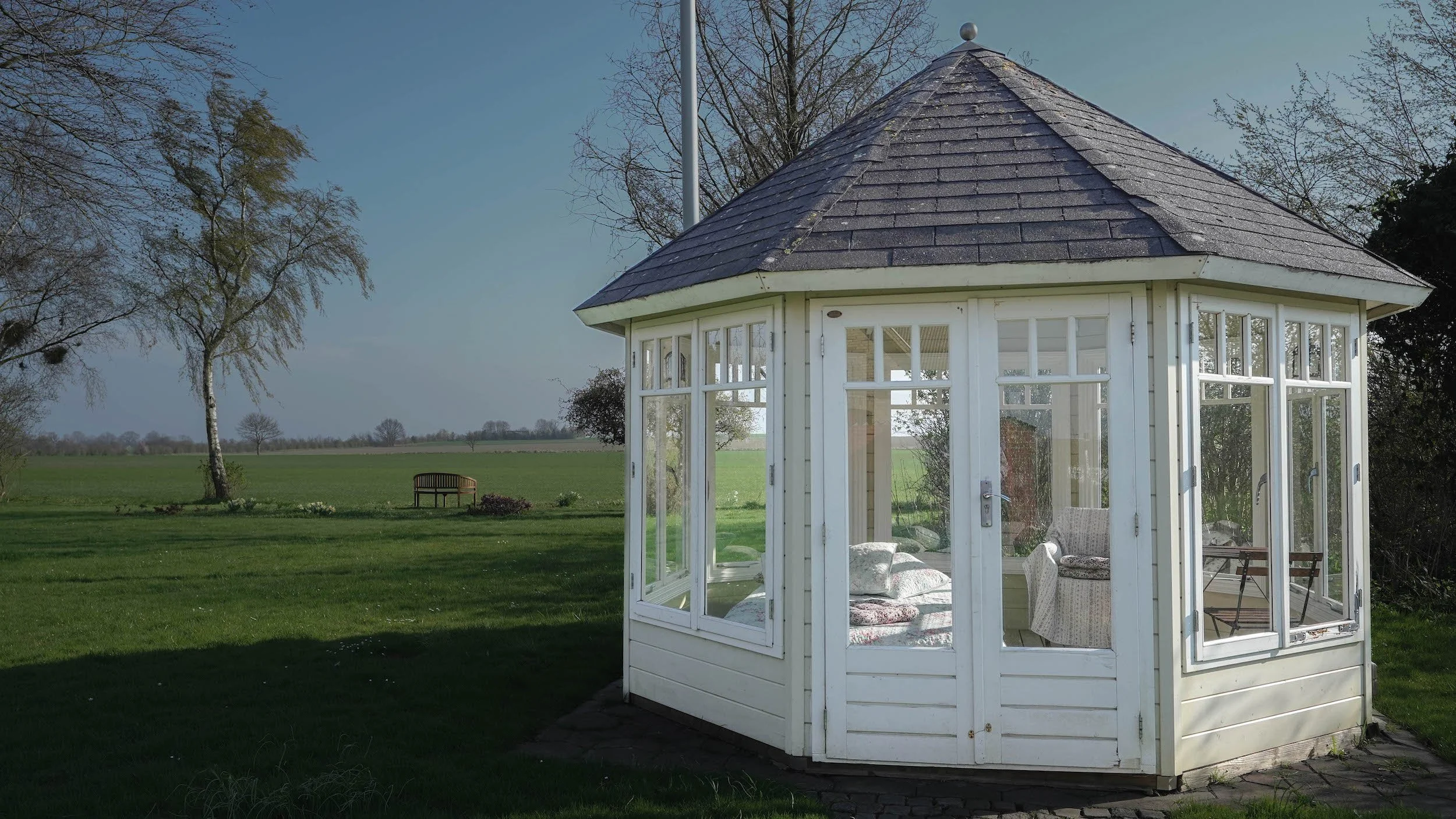 A small white pavilion or gazebo with glass windows and a shingled roof, situated on a grassy field with trees and a bench in the distance.