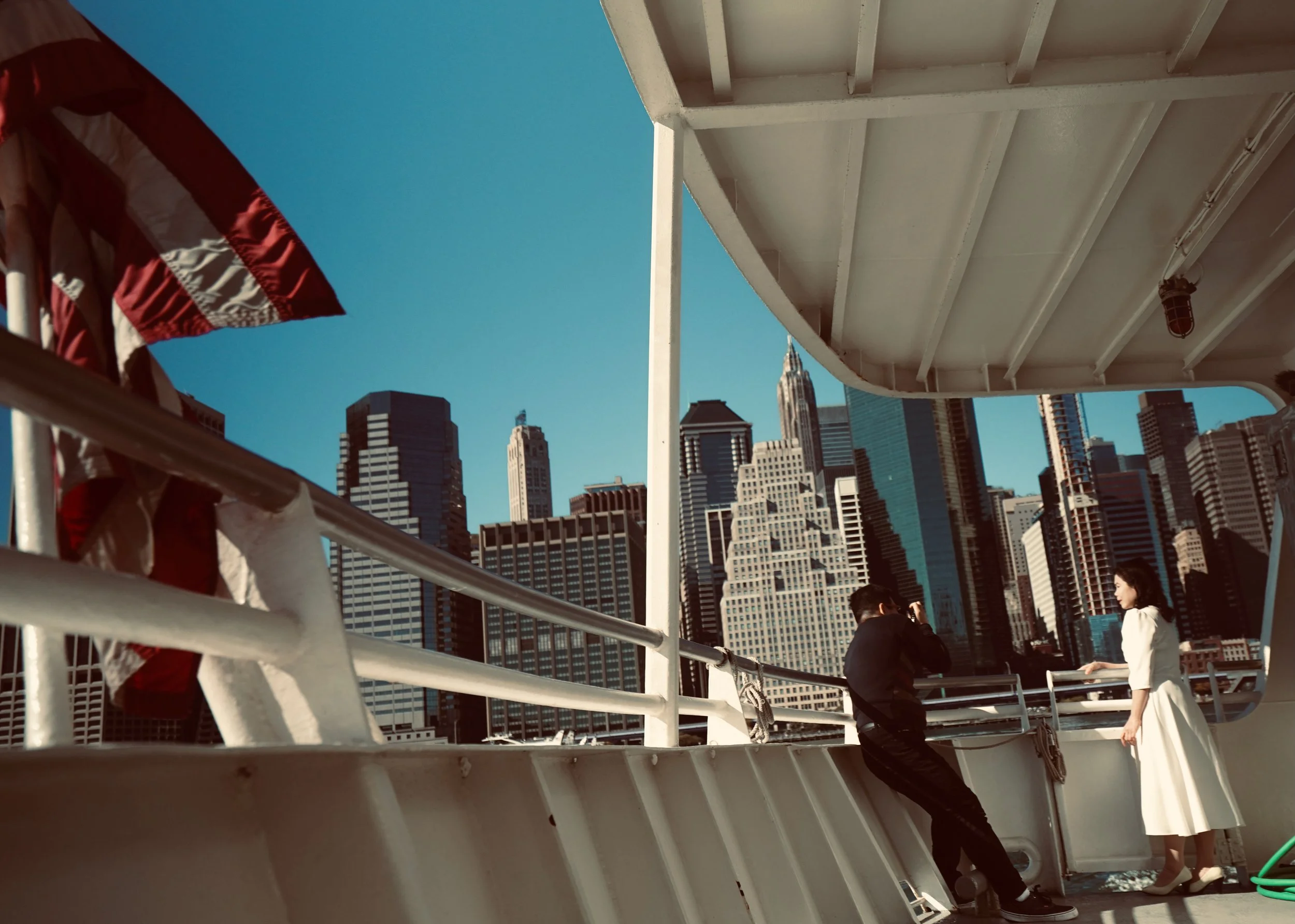 A woman in a white dress and heels is standing on a boat, having her photo taken by a man in black clothing with a camera. In the background, there is a city skyline with tall, modern skyscrapers, and the sky is clear and blue.