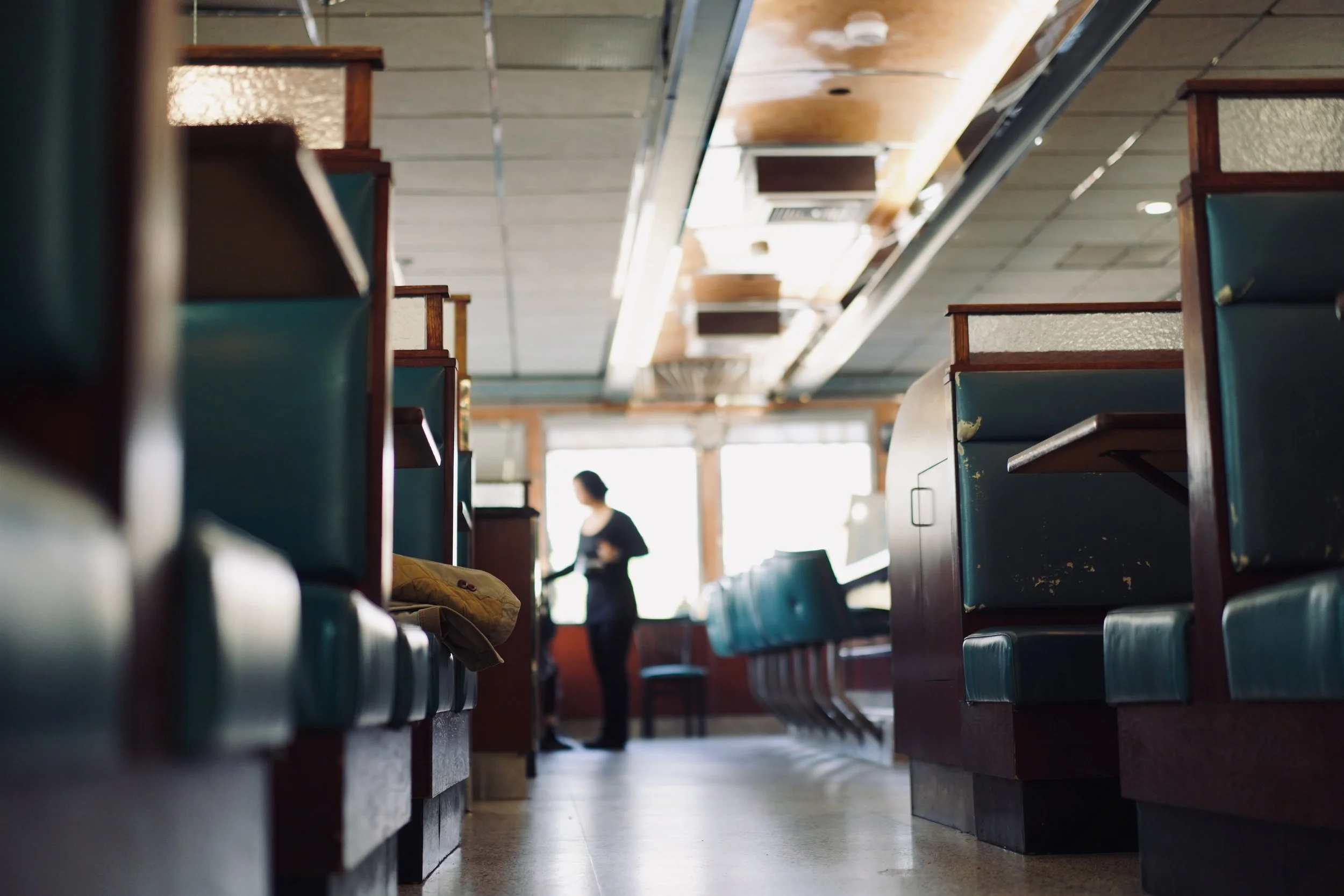 Empty restaurant with teal booths and wooden accents, a person in the background near the counter, illuminated by sunlight through large windows.