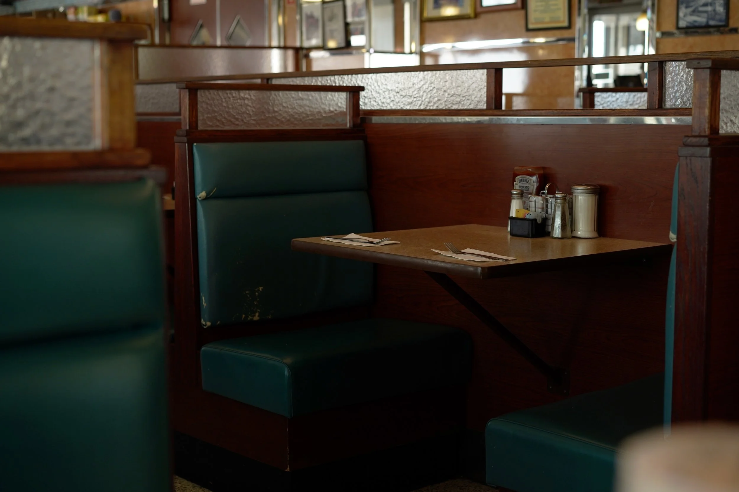 Empty booth in a diner with green leather seats, a wooden table set with napkins, forks, and condiments including ketchup, salt, and pepper shakers.