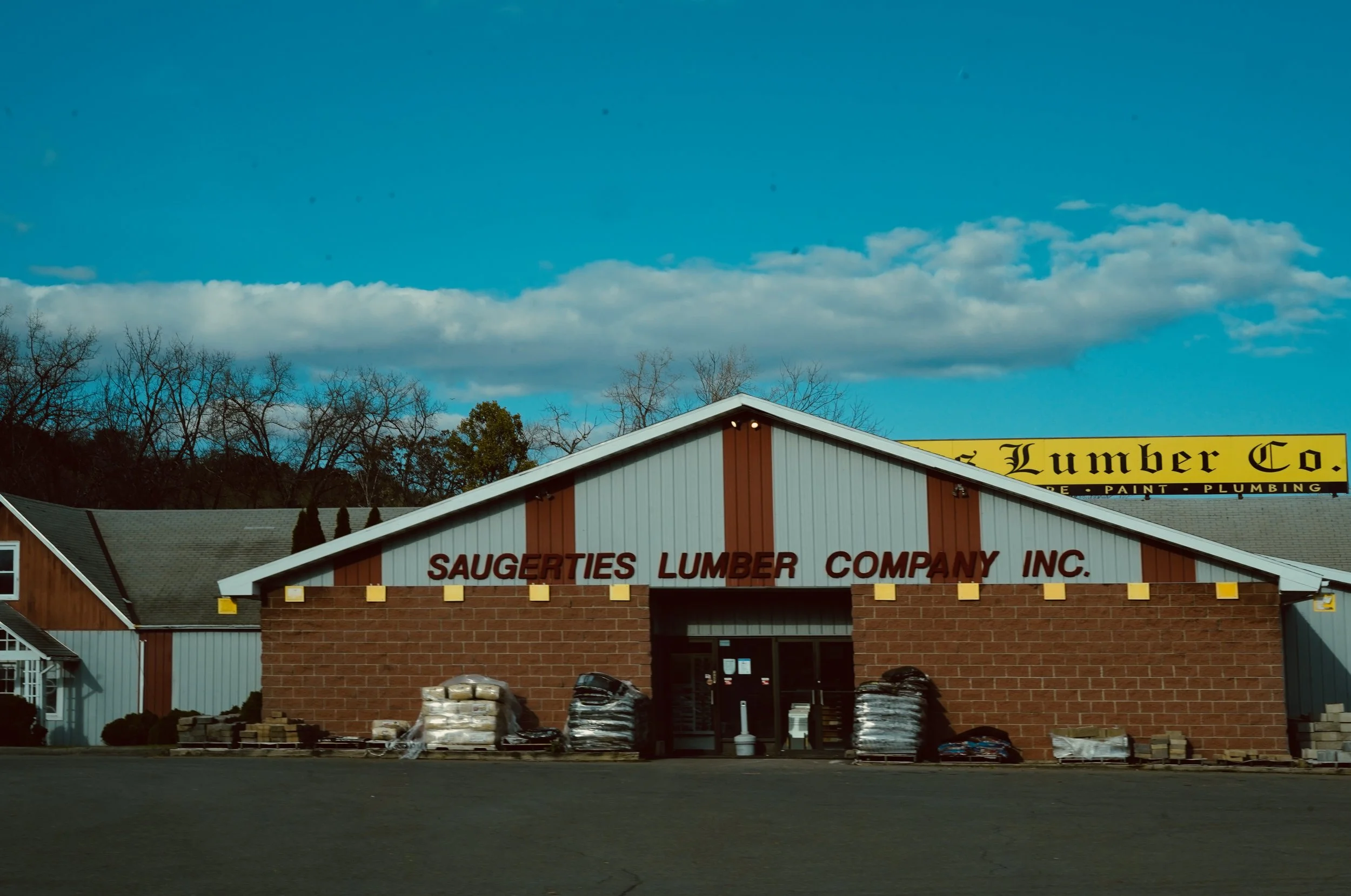 The exterior of Saugerties Lumber Company INC. building with a brick facade and a white roof, with stacks of lumber and building materials outside, under a partly cloudy sky.