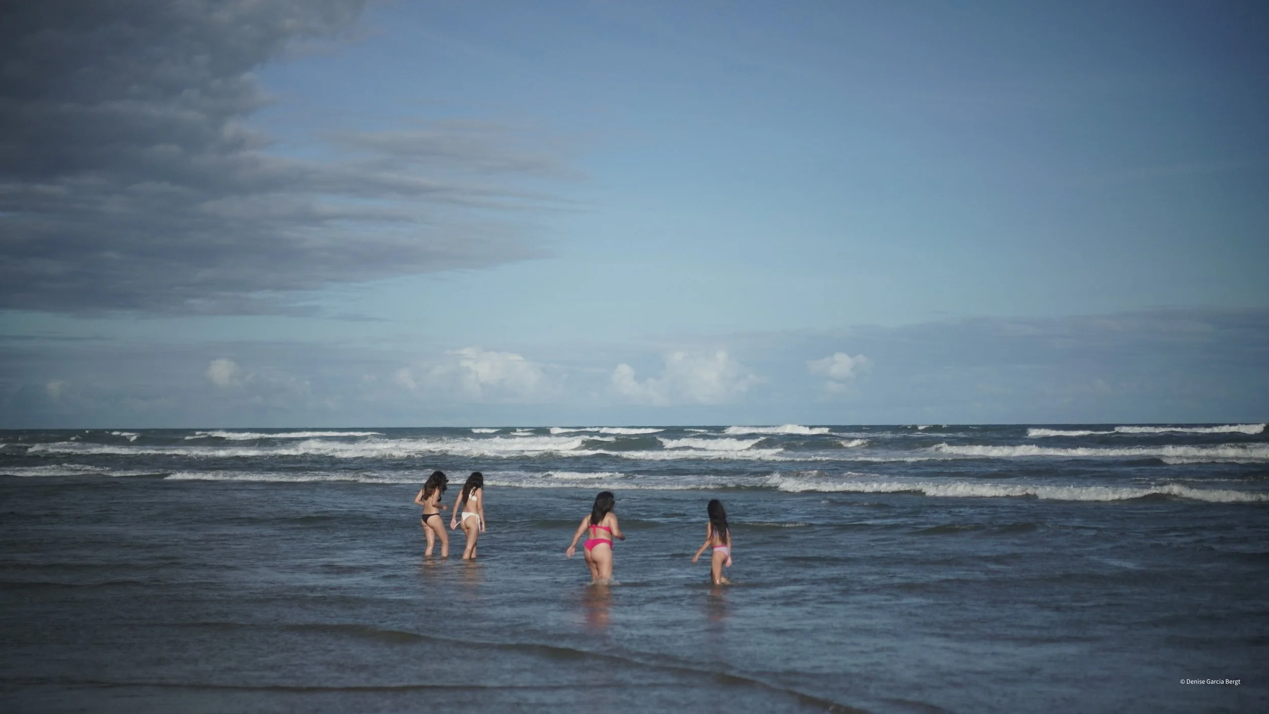 Four girls in swimsuits wading in the shallow ocean water at the beach, with waves and cloudy sky in the background.