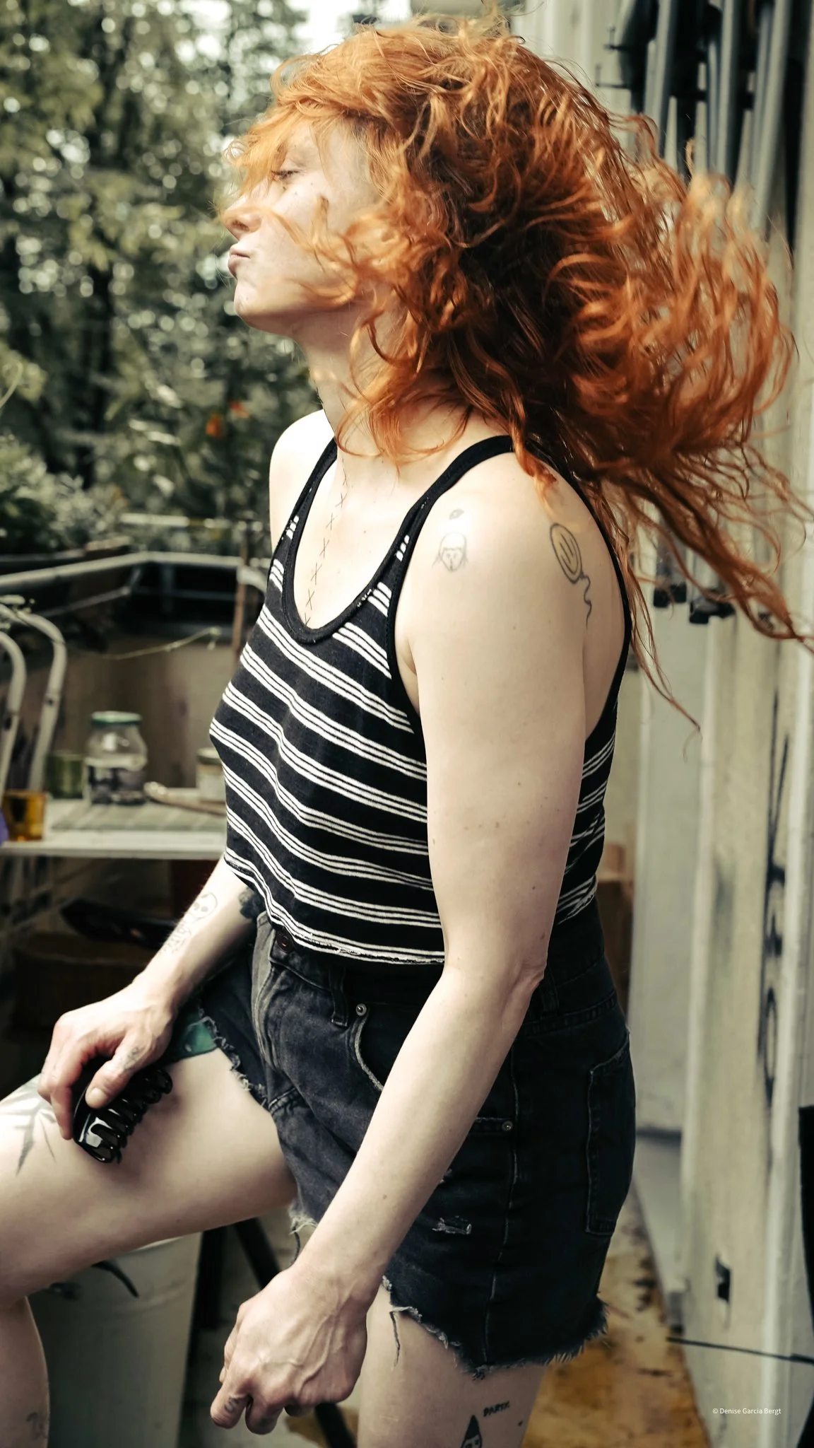A woman with long red hair, wearing a black and white striped tank top and black denim shorts, standing outdoors with trees in the background. She has tattoos on her arms and one on her thigh, and is holding an object in her right hand while flipping