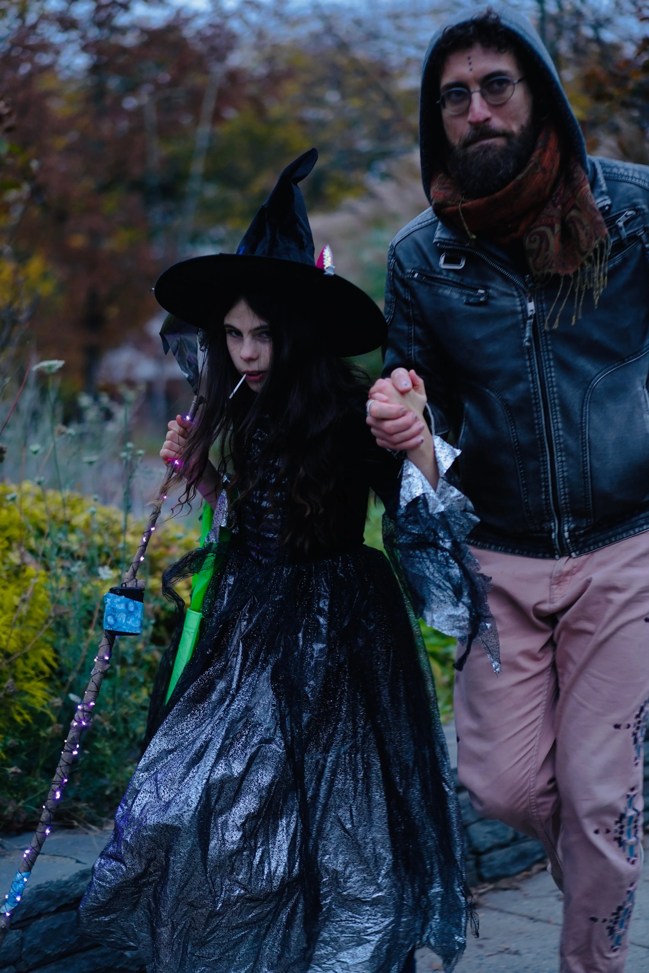 A young girl dressed as a witch with a pointy hat and black dress holding a staff, walking hand-in-hand with an adult man outdoors during fall.