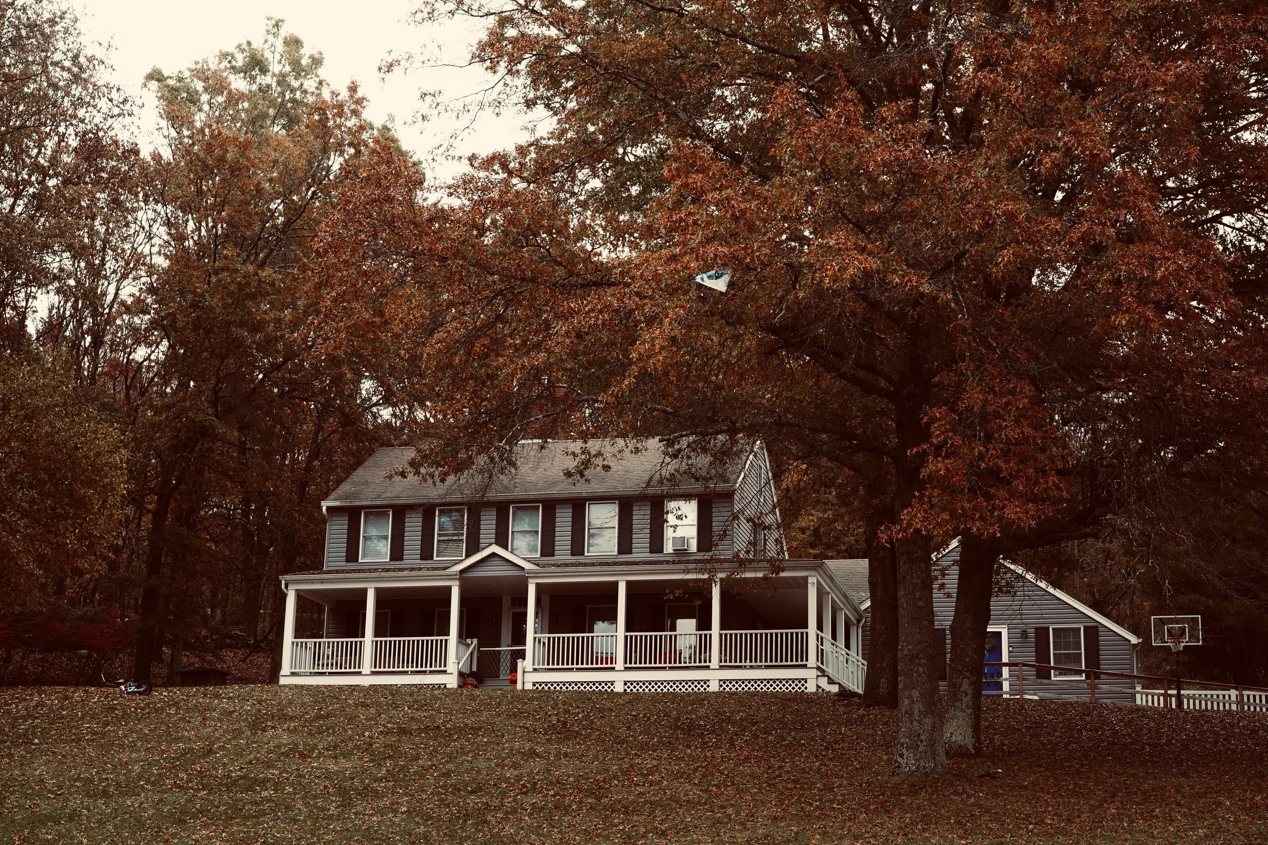 A two-story house with a large porch surrounded by trees with autumn-colored leaves.