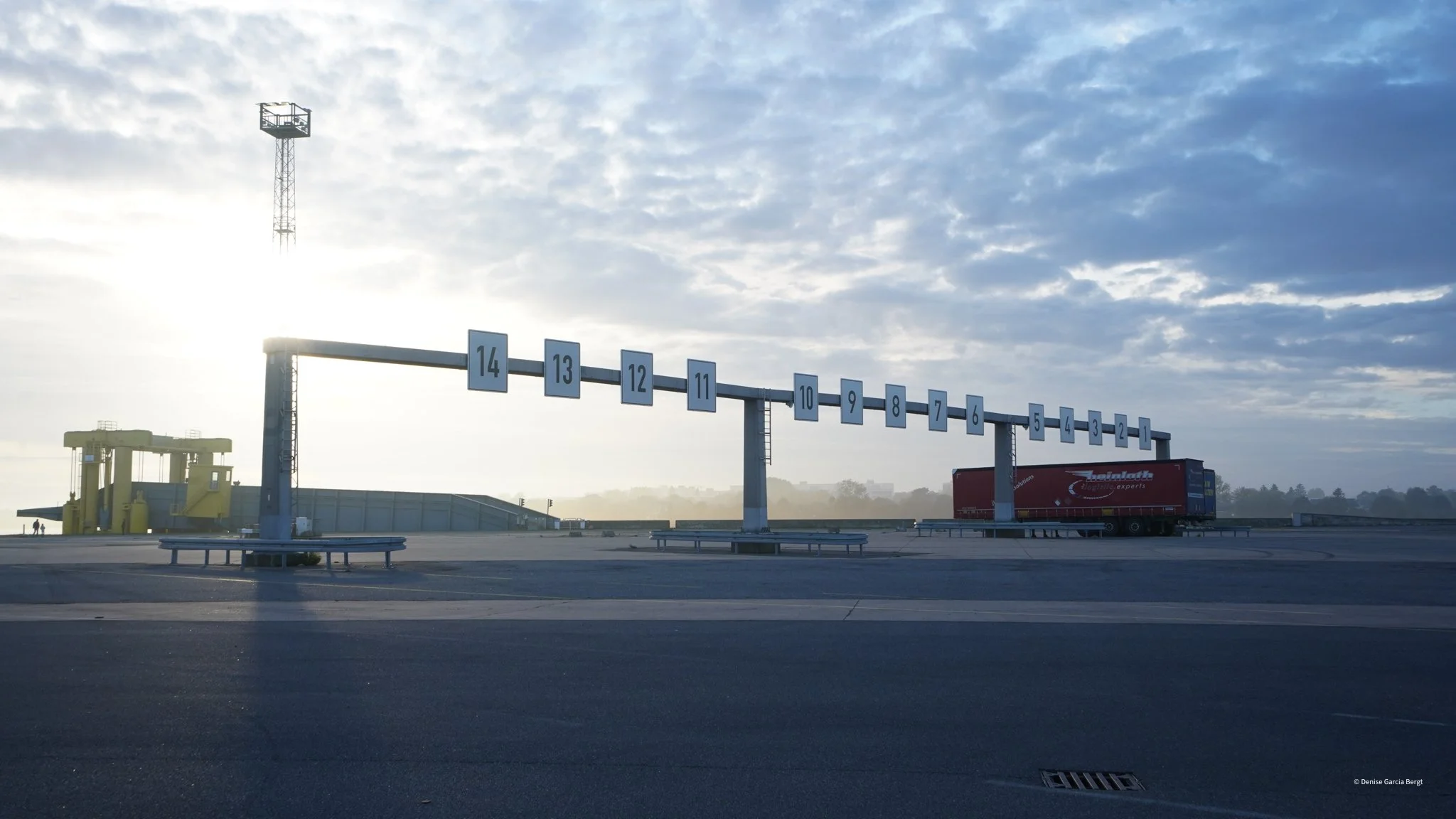 A race track starting grid with number signs from 14 to 1, a yellow vehicle, a red truck, and a tall light tower under a cloudy sky.