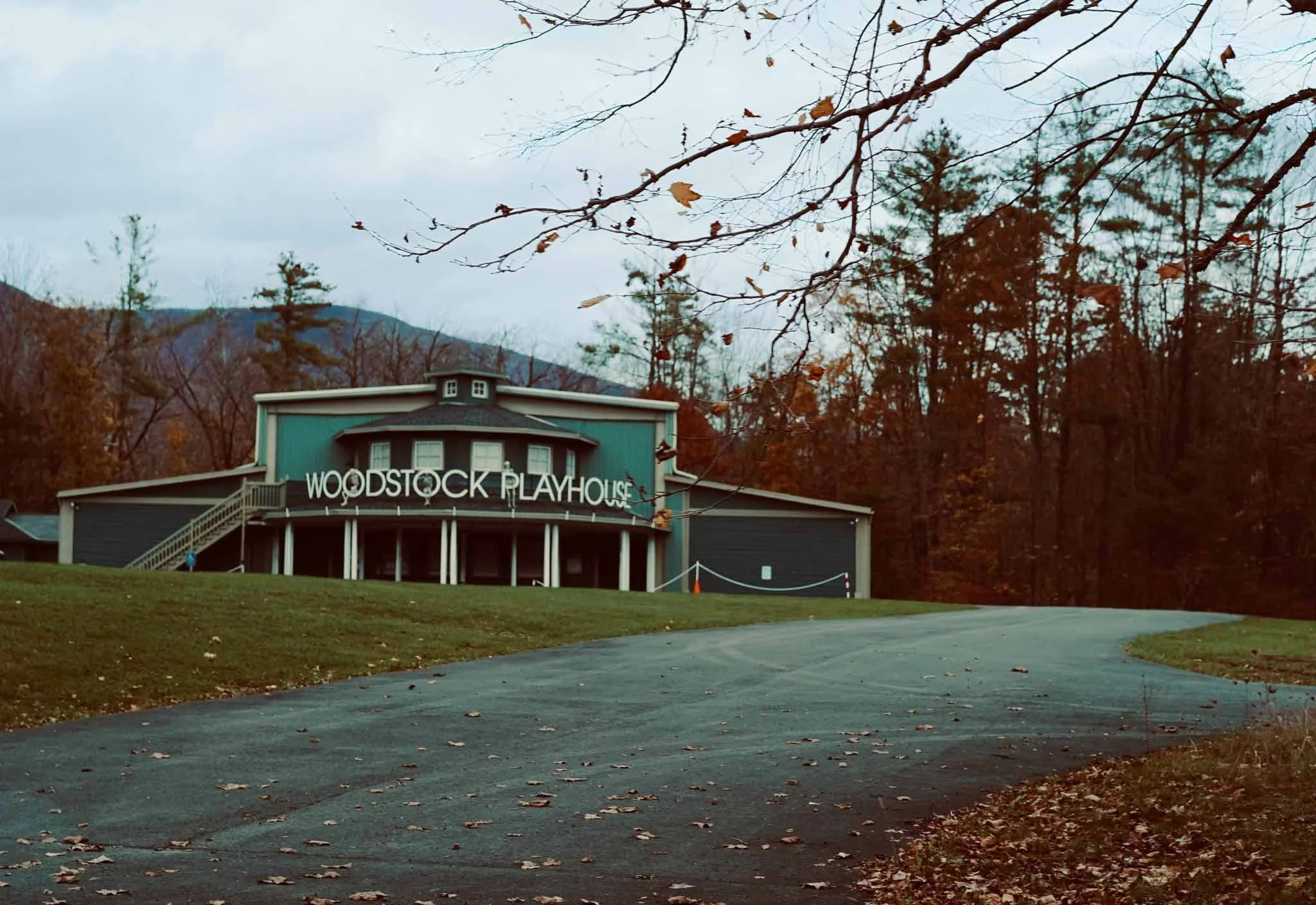 Woodstock Playhouse, a dark green and blue building with a curved roof and white sign, situated on a grassy area with fallen leaves, surrounded by leafless trees and mountains in the background on a cloudy day.