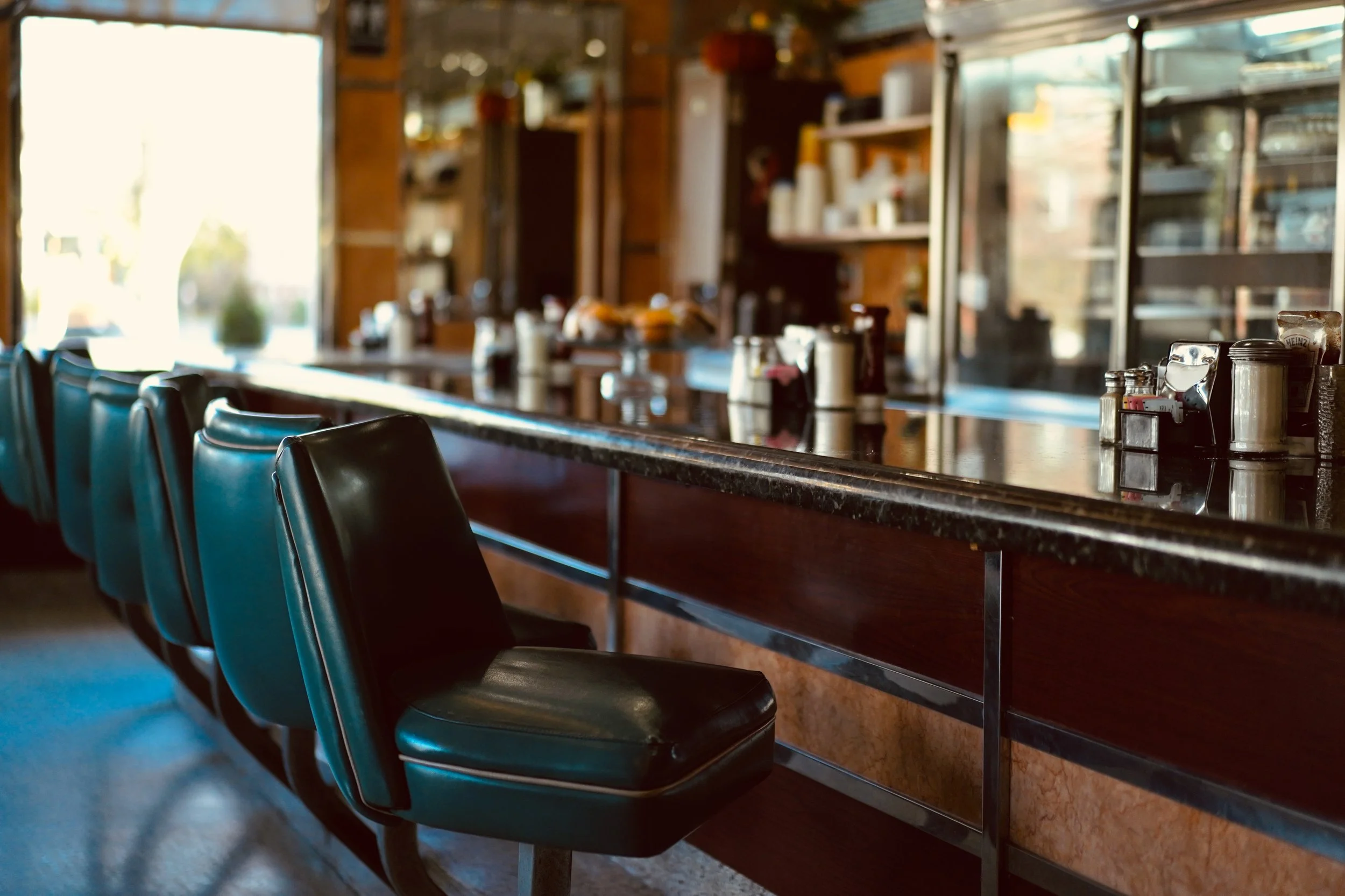 Empty retro-style diner with black vinyl bar stools aligned along a dark granite counter, condiments and utensils on the counter, and warm sunlight streaming through the large window.