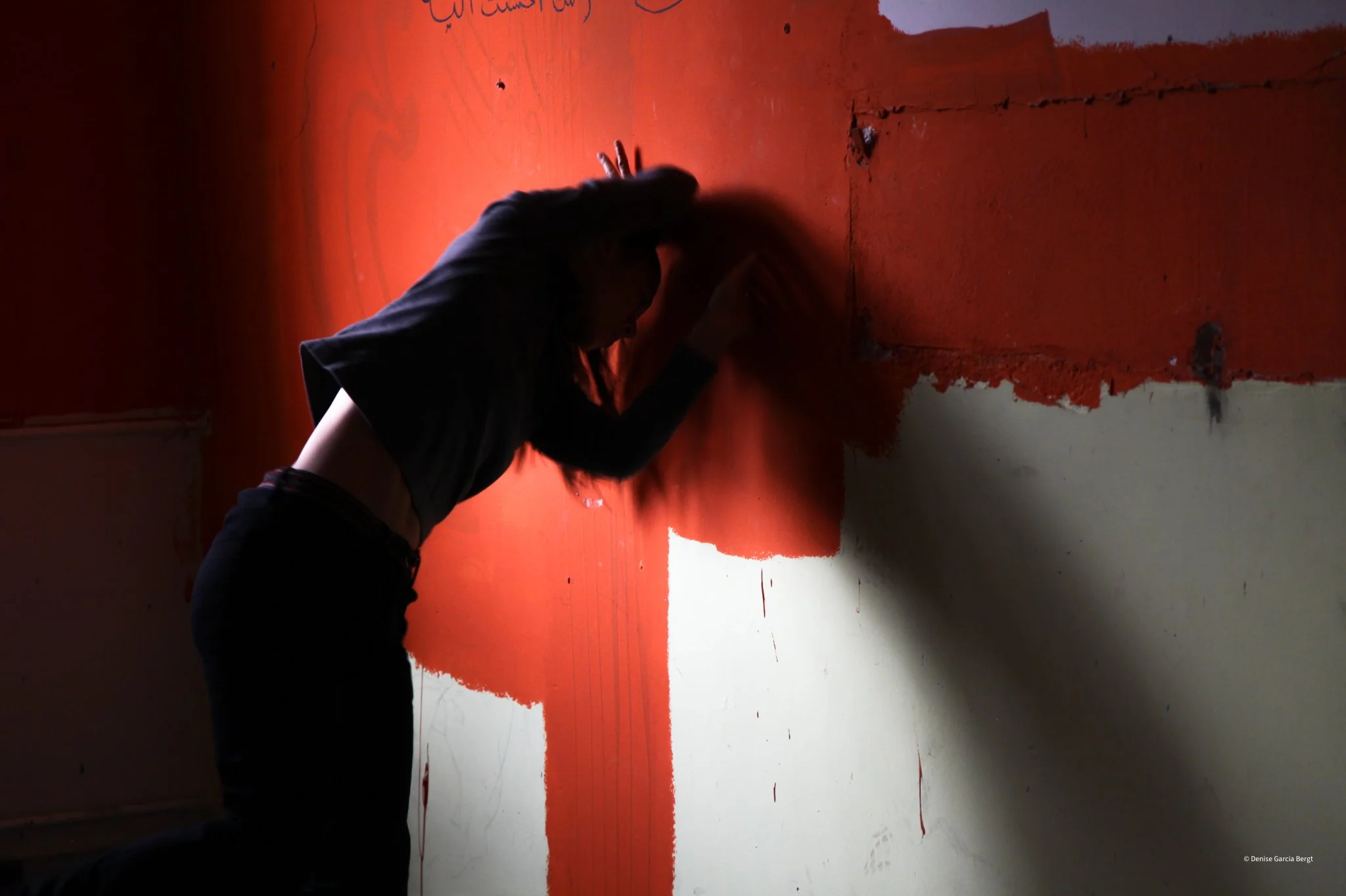 A person with long hair is working on a wall, painting it with red paint. The person is leaning against the wall with one hand on the wall and the other arm bent. The wall has a large section painted red, with some areas still unpainted or partially 