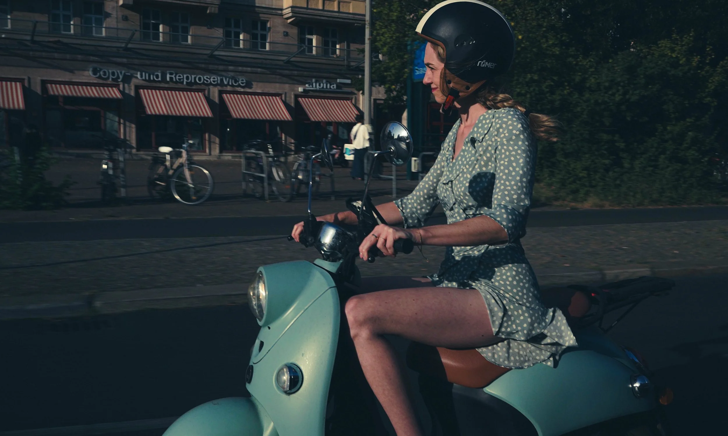 Woman riding a light blue scooter on a city street, wearing a black helmet and a green dress with white polka dots, during daytime.